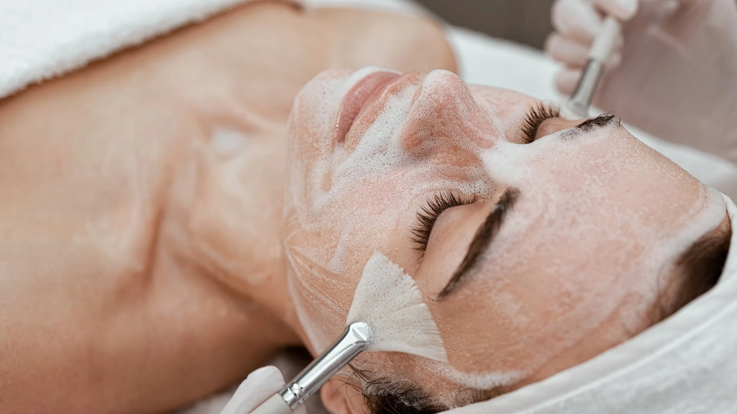 A woman is lying on a treatment bed with soap foam on her face, receiving a facial treatment from a professional using a device.