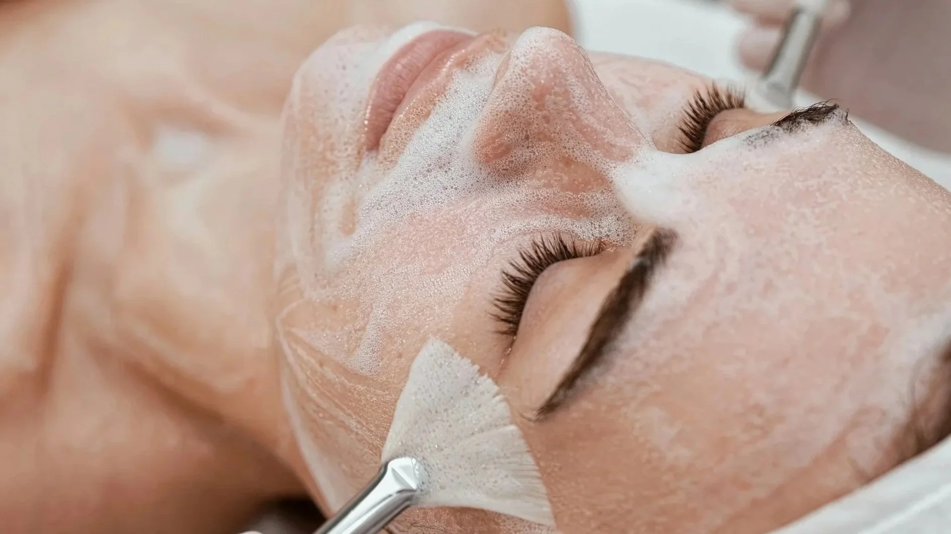 Close-up of a woman with closed eyes receiving a facial treatment, with foam on her face, at a spa or beauty clinic.