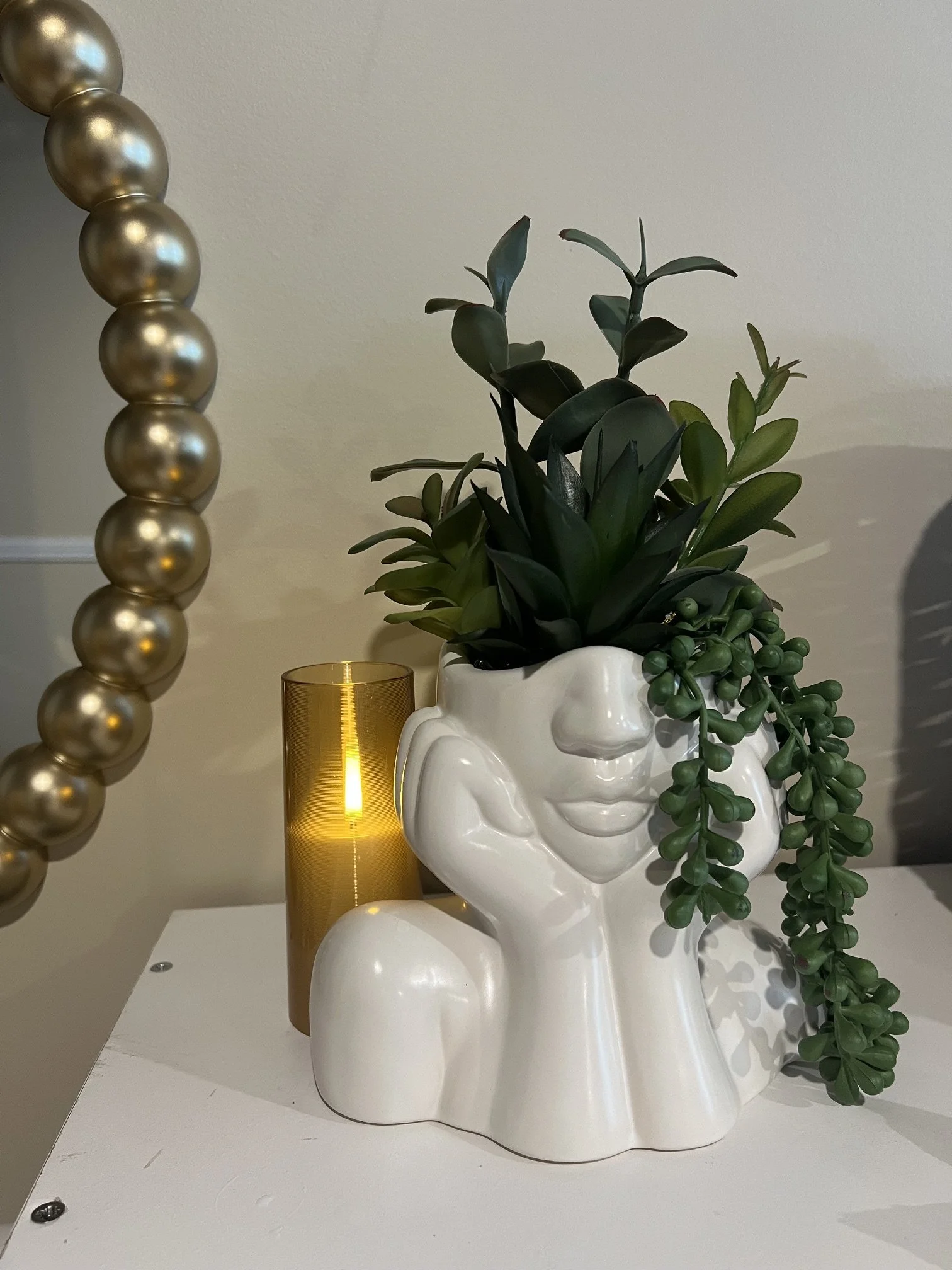 Decorative arrangement with a potted green succulent plant in a white ceramic skull-shaped vase, a gold candle holder with a candle, and a gold beaded ornament on a white surface against a beige wall.