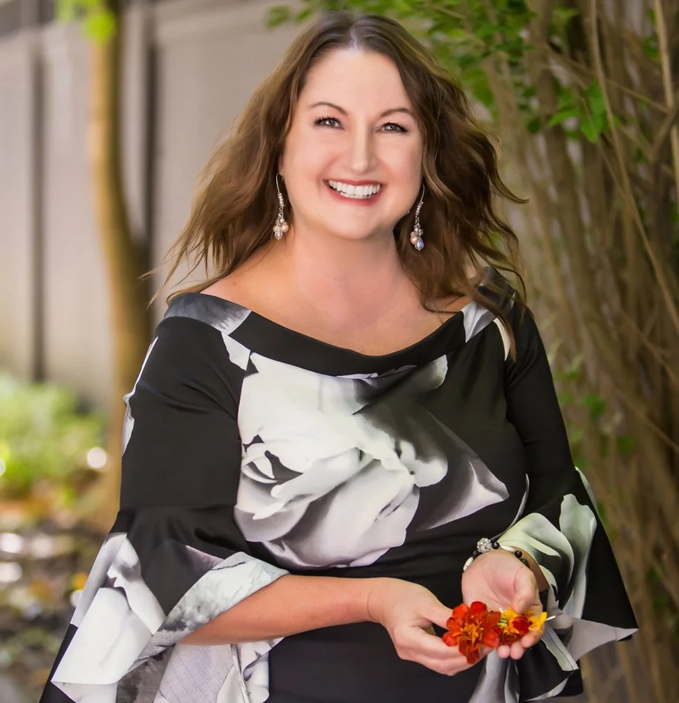 A cheerful woman with long brown hair, wearing a black and white floral dress and earrings, holding a few yellow and red flowers outdoors with greenery and a fence in the background.