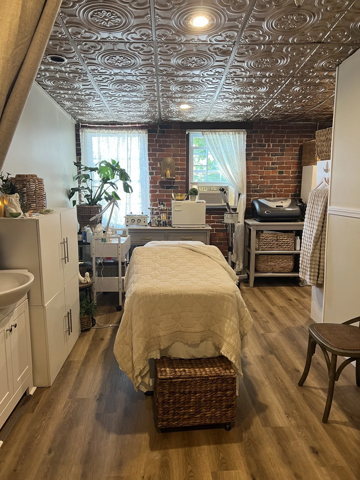 A cozy room with wood flooring and an exposed brick wall. There is a bed with a beige quilt in the center, a woven basket at the foot, and a small rolling cart with medical or skincare equipment. The room has a white cabinet, a potted plant, and natural light coming through two windows with sheer curtains. The ceiling has ornate metal tiles and recessed lighting.