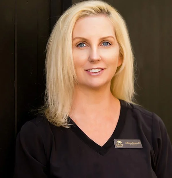 A blonde woman smiling, wearing a black top with a name tag, standing outdoors.