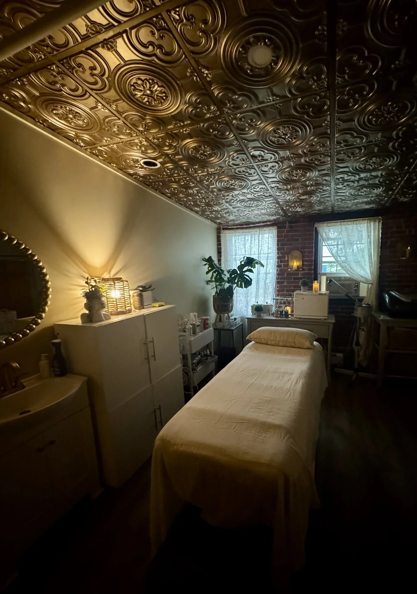 A dimly lit room with a decorative tin ceiling, a bed with a yellow bedspread, a white dresser with a mirror, plants, and various small items near the windows with sheer curtains.