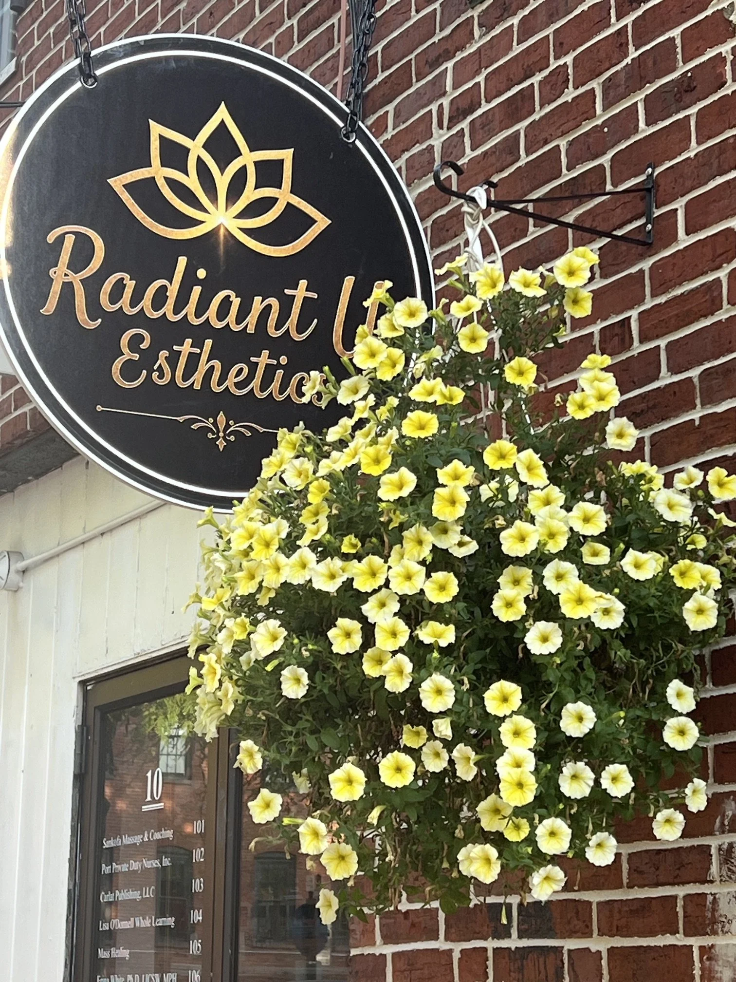 A black round sign with gold and white text that says 'Radiant' and 'Esthetic' above a stylized gold lotus flower logo, hanging on a brick wall next to a window with a yellow flower arrangement in front.