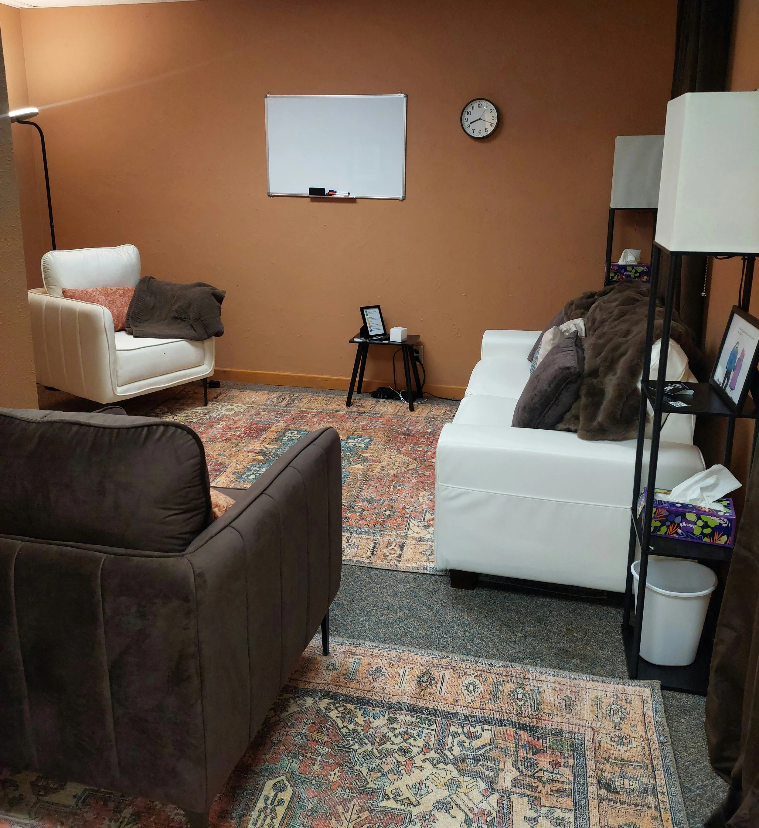 A cozy living room with beige walls and an oriental rug. There are white and brown sofas with cushions, a small black side table with a tablet device and a speaker, a whiteboard on the wall, a clock showing 2:45, and a tall floor lamp. A shelf with tissue box and framed picture is also visible.