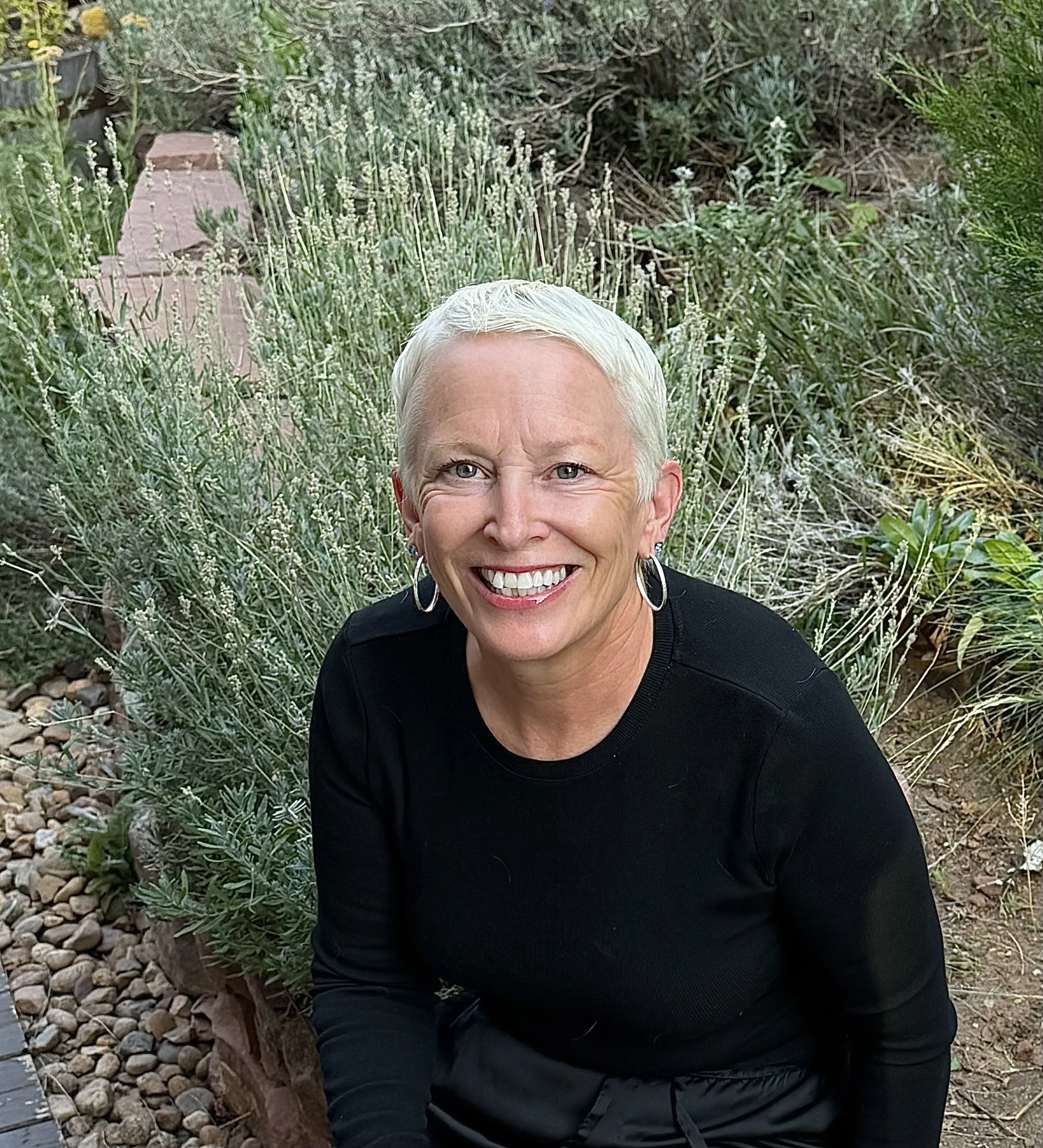 Smiling woman with short blonde hair and earrings, wearing a black top, outdoors among lush green plants and flowers, with a curved brick border and rocky ground nearby