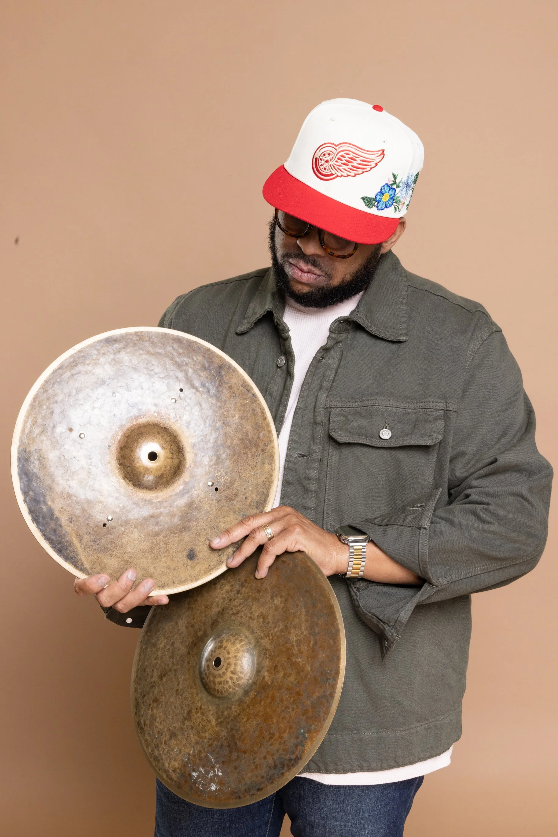 A man wearing glasses, a green jacket, and a white shirt holding up multiple cymbals, with a tan wall in the background.