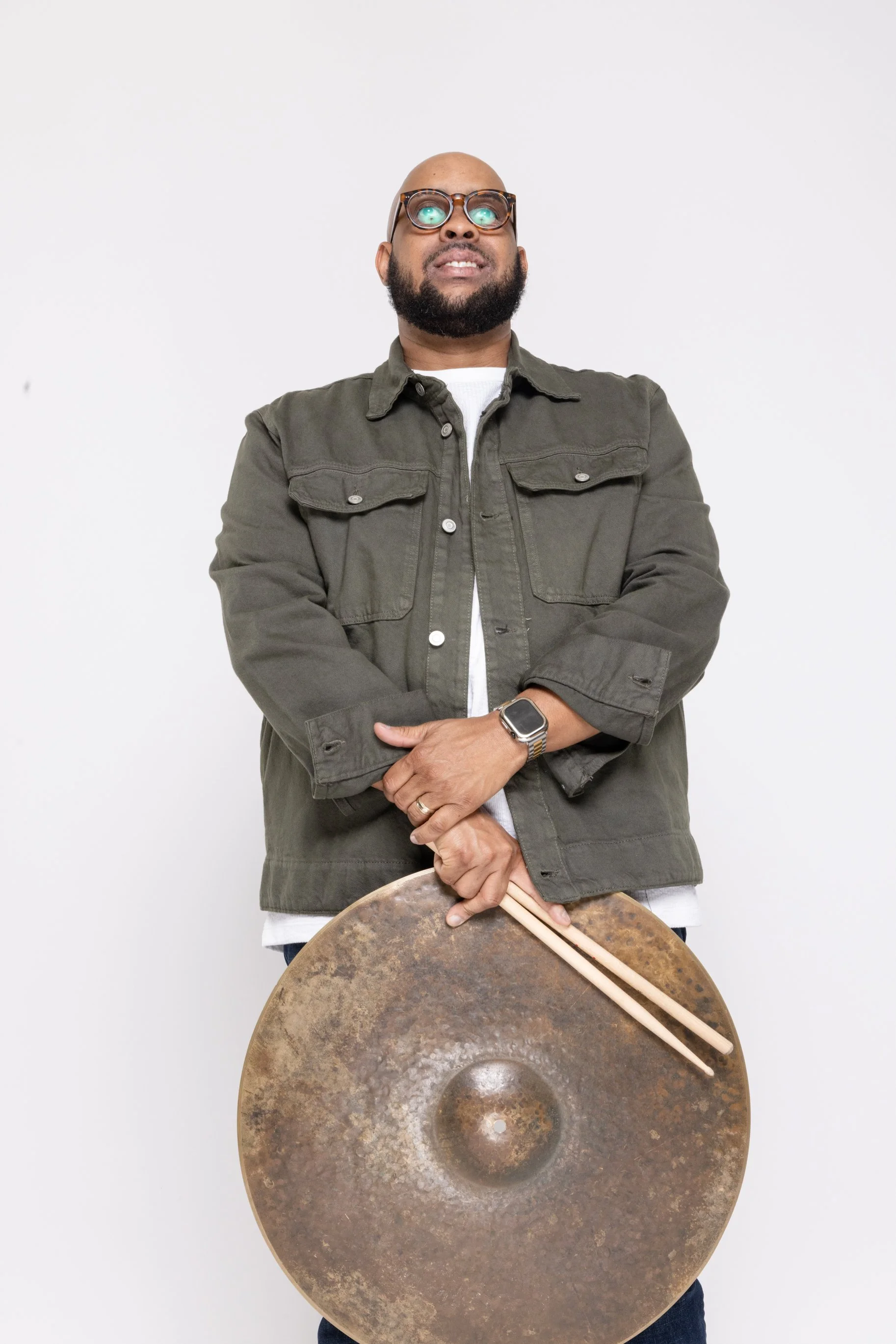 Man with a beard and glasses holding a large metallic gong and wooden mallets, standing against a plain white wall.
