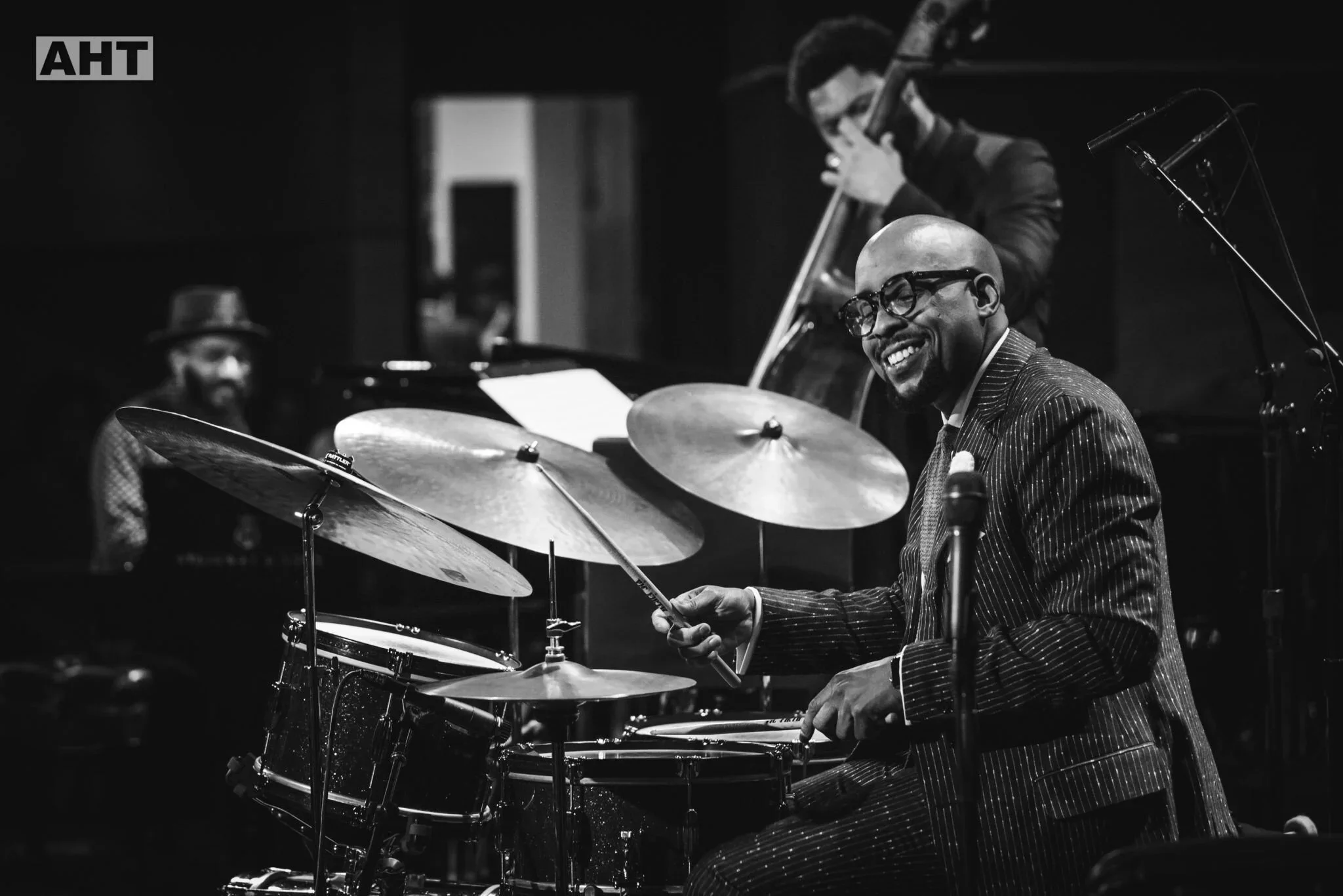 B&W photo of the Henry Conerway trio, featuring a smiling Conerway at the drums in a pinstripe suit and glasses in the foreground, pianist Kenny Banks Jr., partially visible in the left background, and bassist Barry Stephenson in the back. AHT Photo