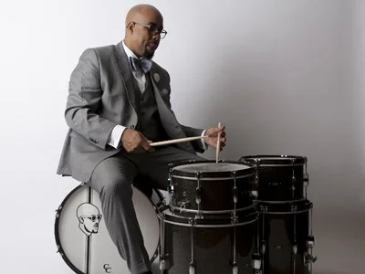 Henry Conerway III in a gray suit playing a snare drum while sitting on his bass drum featuring his logo; a silhouette of his bald head, glasses and goatee, against a plain gray background.