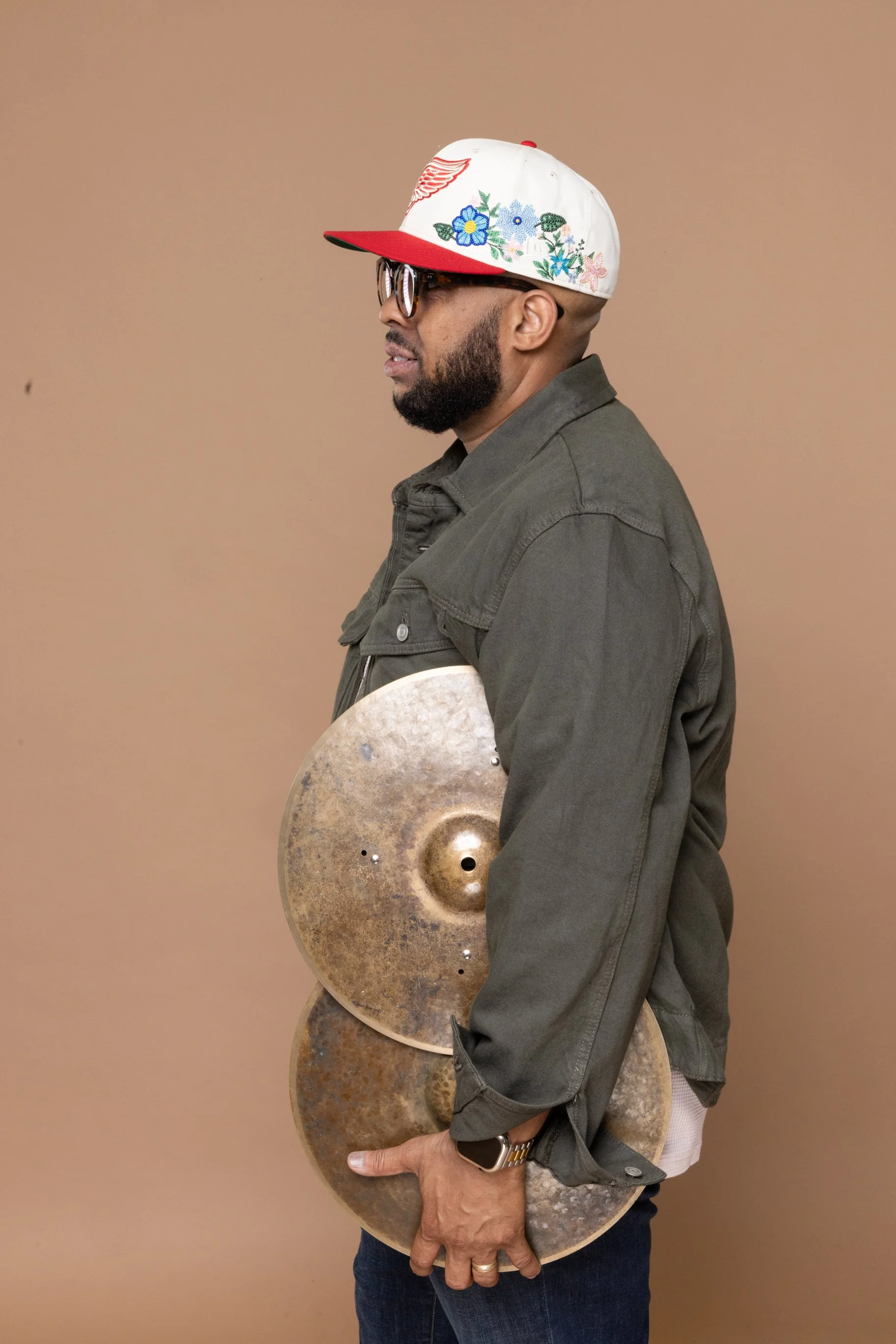 A man wearing a gray jacket with floral embroidery, sunglasses, and a colorful cap with embroidery, holding a pair of rusted cymbals against a beige background.