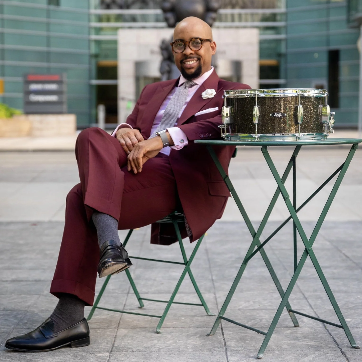 Henry Conerway III in a maroon suit sitting at a green outdoor table in Detroit's Campus Martius with his snare drum on it, smiling at the camera.