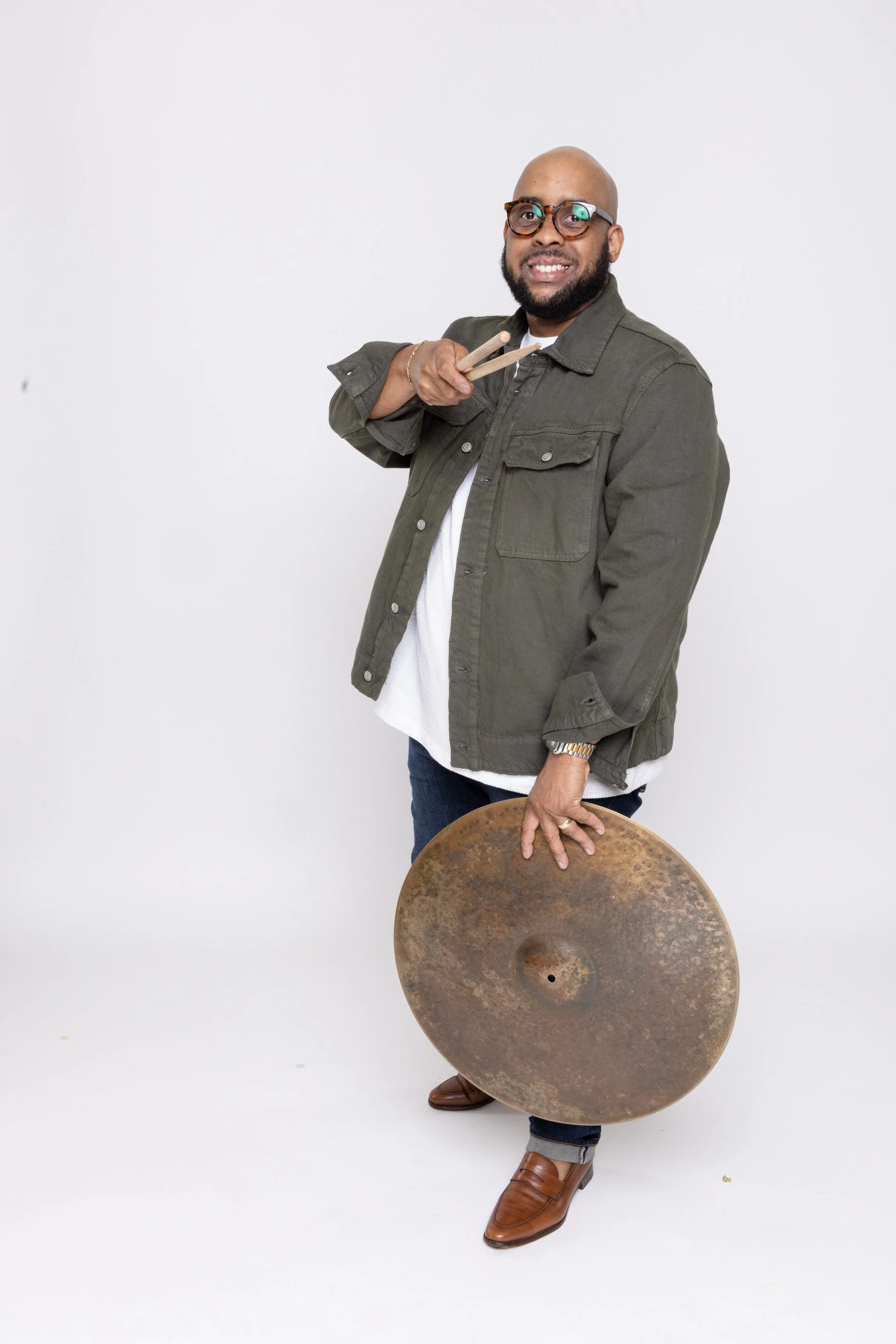 Man with beard and glasses holding drumsticks, standing behind a large, rusty cymbal, wearing a green jacket, white shirt, dark jeans, and brown shoes, against a plain white background.