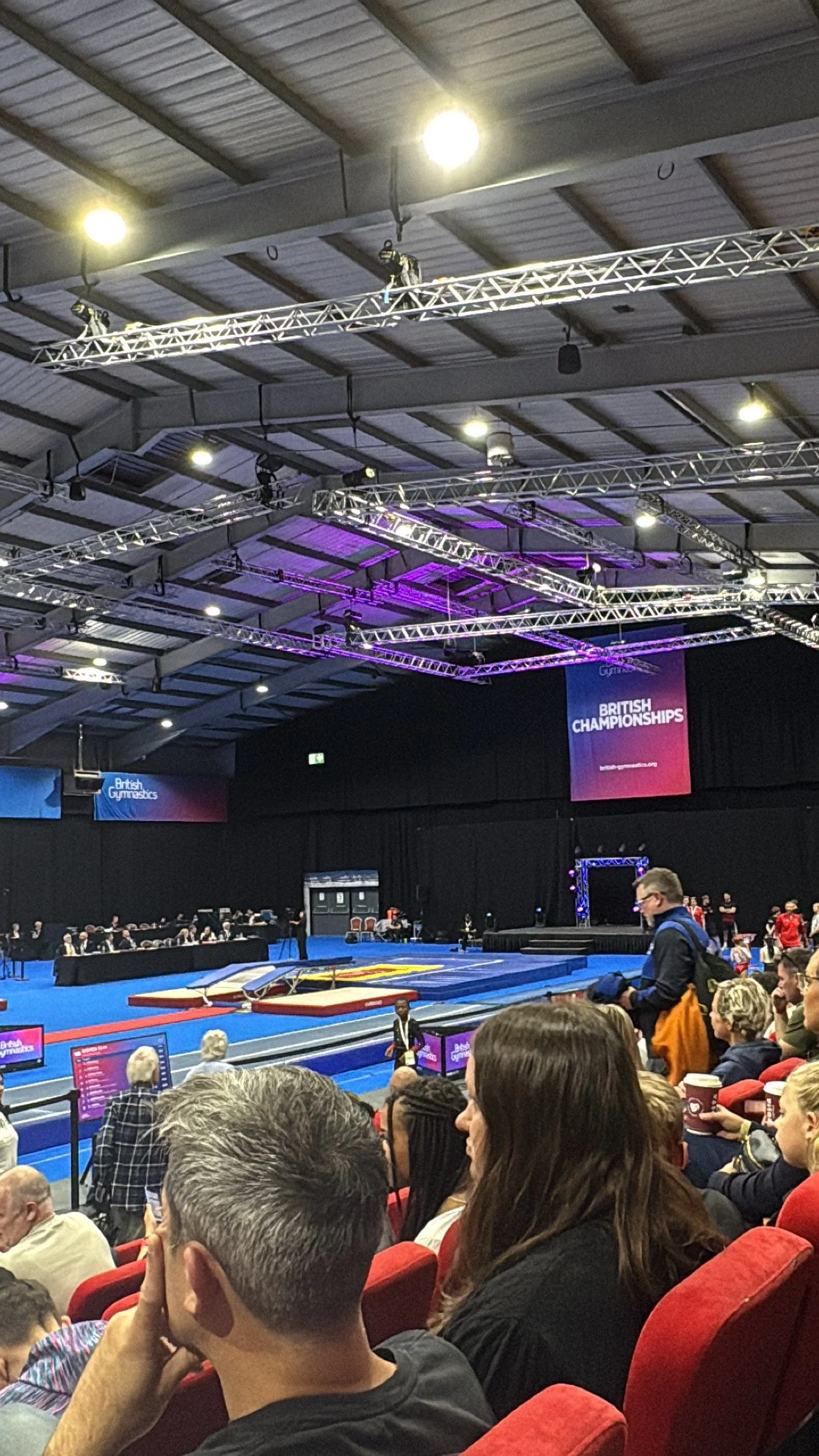 Indoor gymnastics arena with competitive event setup, audience seated in red chairs, banners display 'British Championships,' multiple judges and gymnasts preparing.