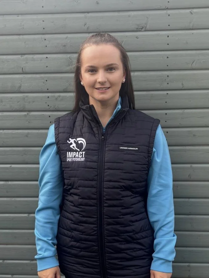 Young woman standing in front of a gray wooden wall, wearing a black Impact Sport Psychology jacket with blue sleeves.