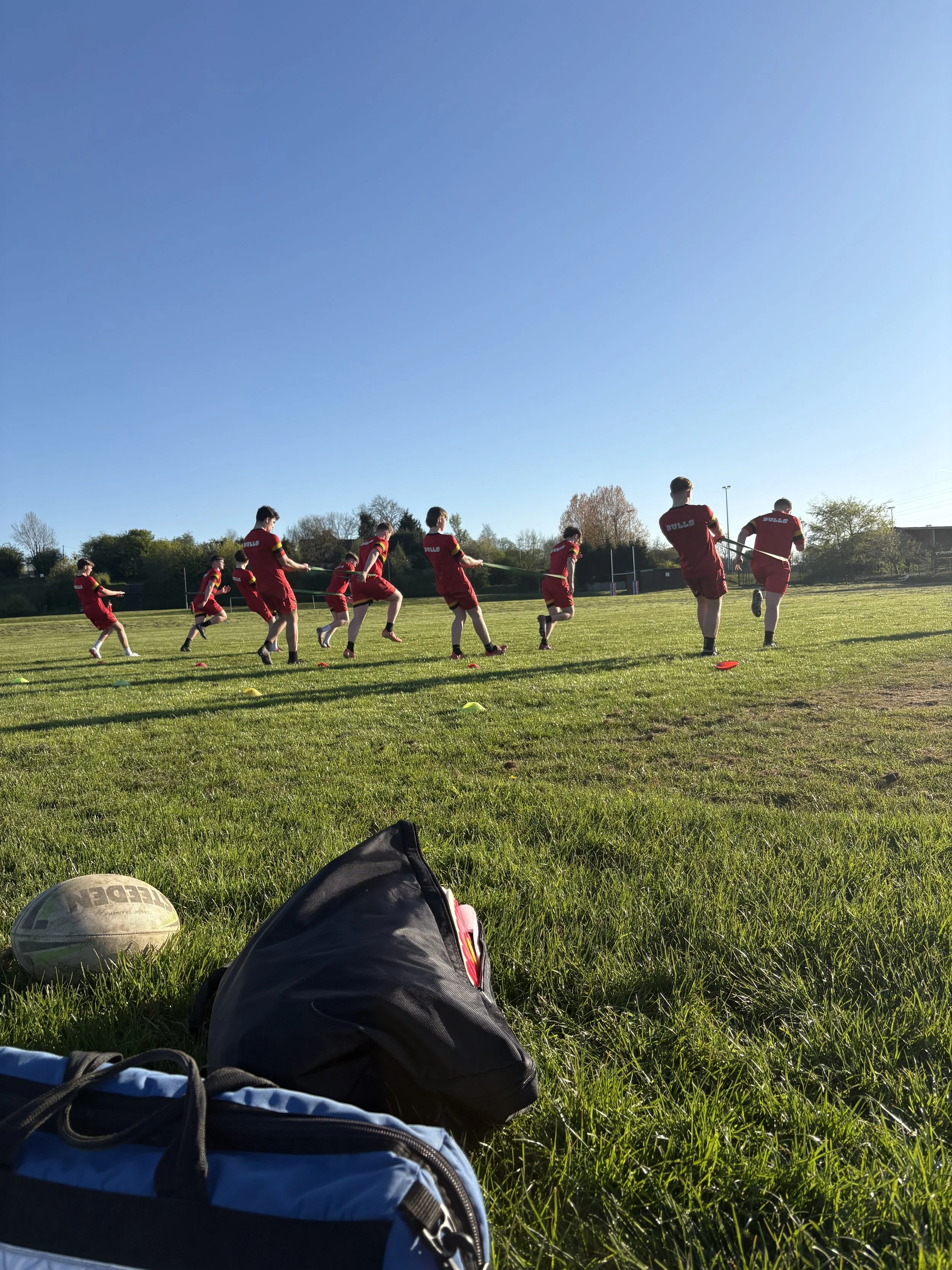 A group of young athletes wearing red shirts and shorts are running on a grassy field during daytime. The scene is captured from a low angle, with sports bags and a rugby ball in the foreground, and trees and a clear blue sky in the background.