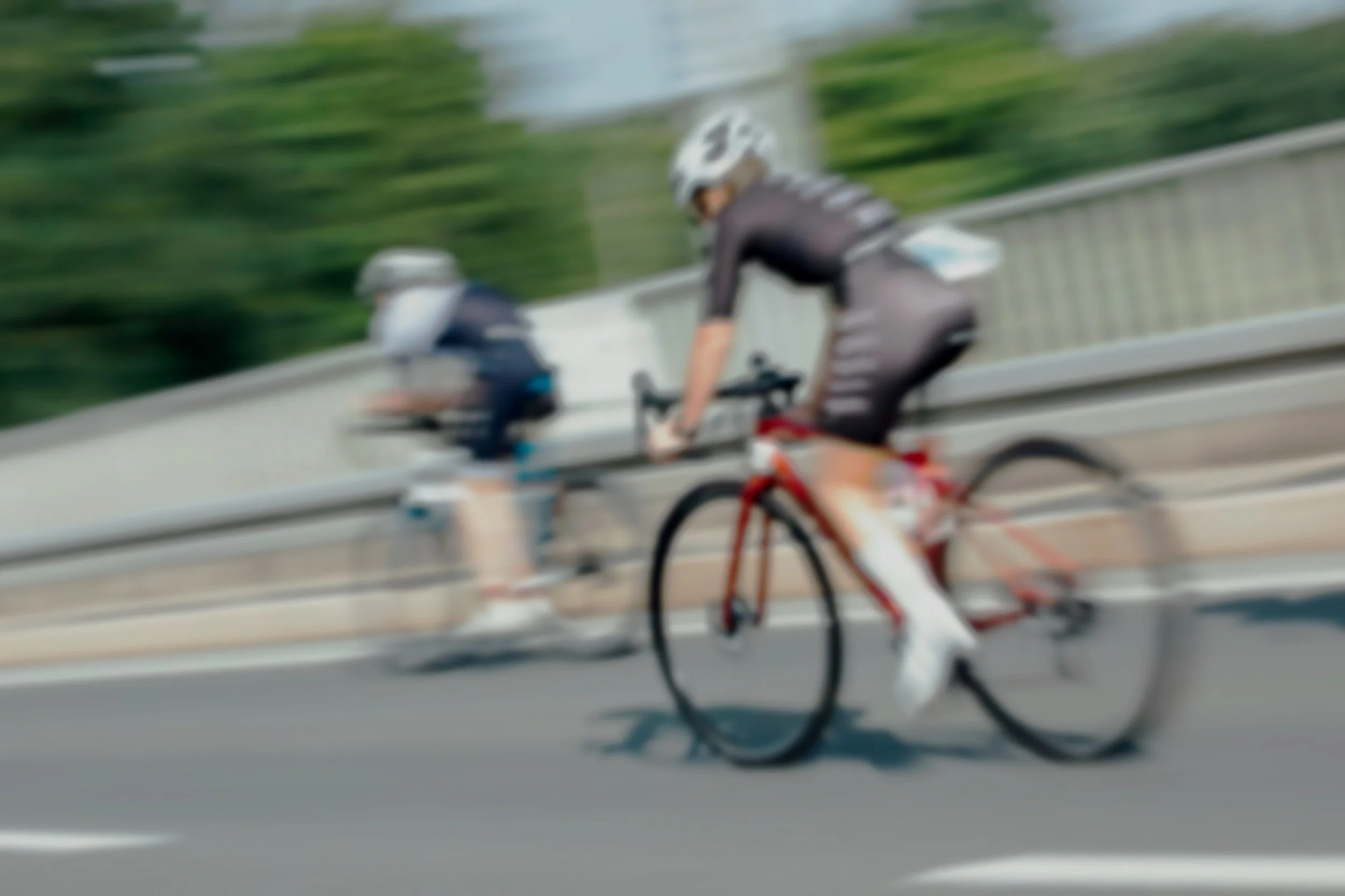 Blurred image of two cyclists riding on a roadway, viewed from a moving vehicle, with trees and greenery in the background.