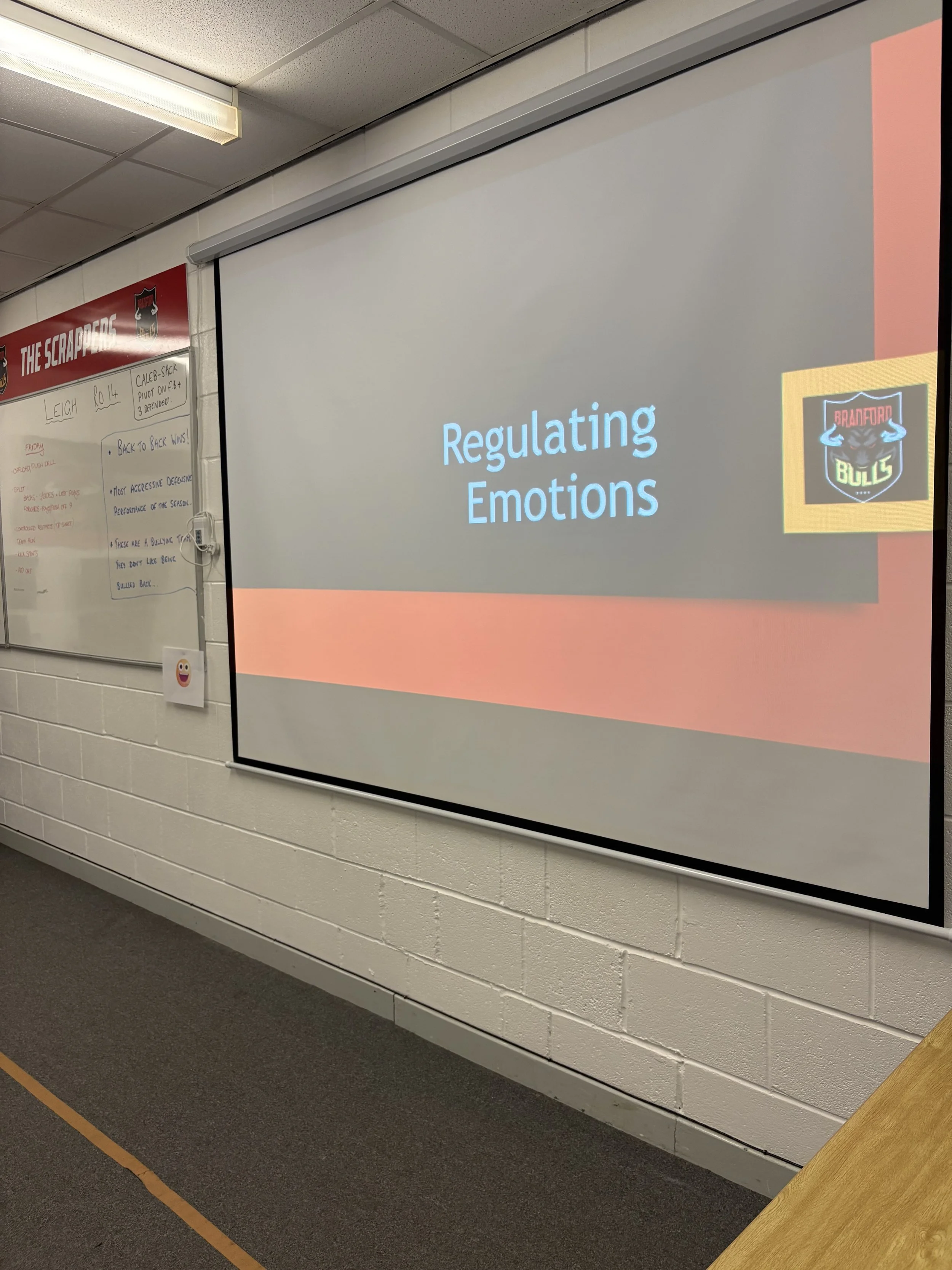 Classroom projector screen displaying a presentation slide titled 'Regulating Emotions' located at the front of a classroom. Next to the screen, a whiteboard has notes and a logo of the Bradford Bulls rugby team.