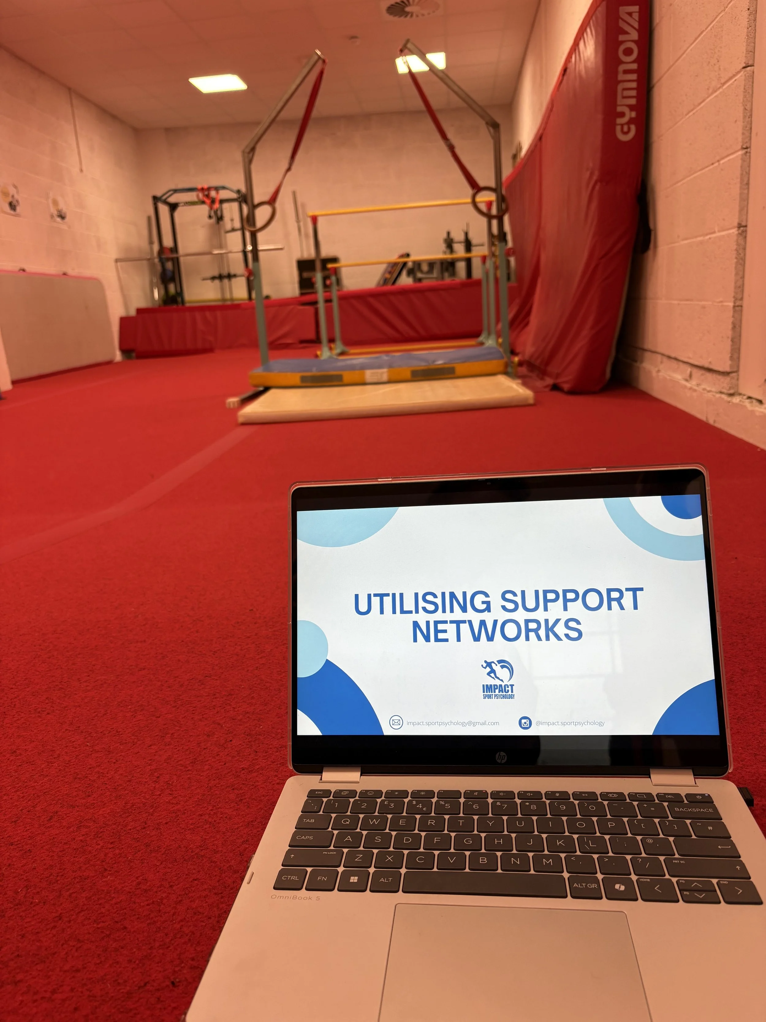 A laptop on a red carpeted surface displaying a presentation slide titled 'Using Support Networks' with impact sports psychology logo and contact information. In the background, gymnastic equipment including rings and mats are visible in a room with 