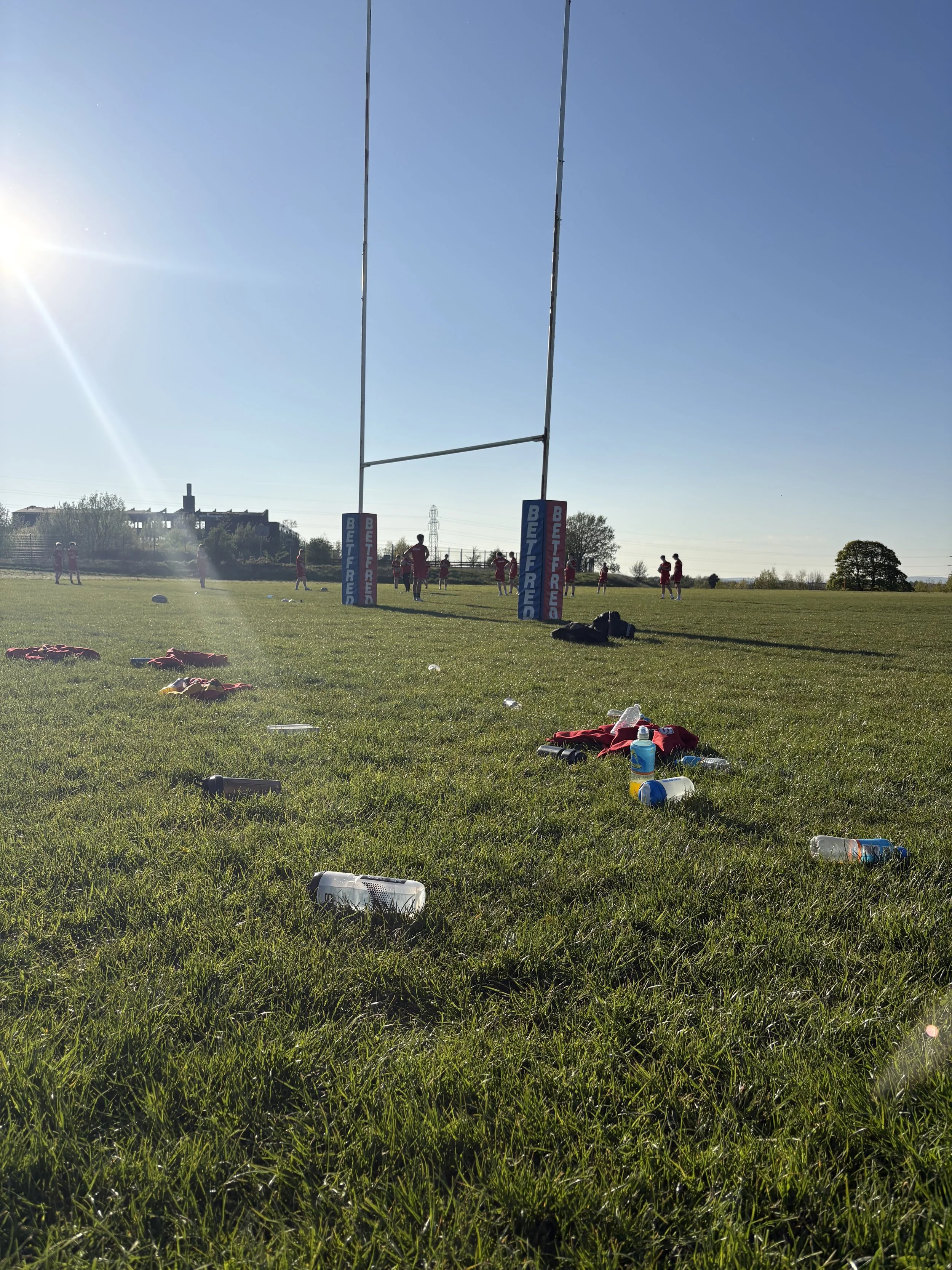 A sports field with a rugby goalpost and a group of athletes practicing nearby. The ground is littered with water bottles, clothing, and other items. The sun is shining brightly in a clear blue sky.