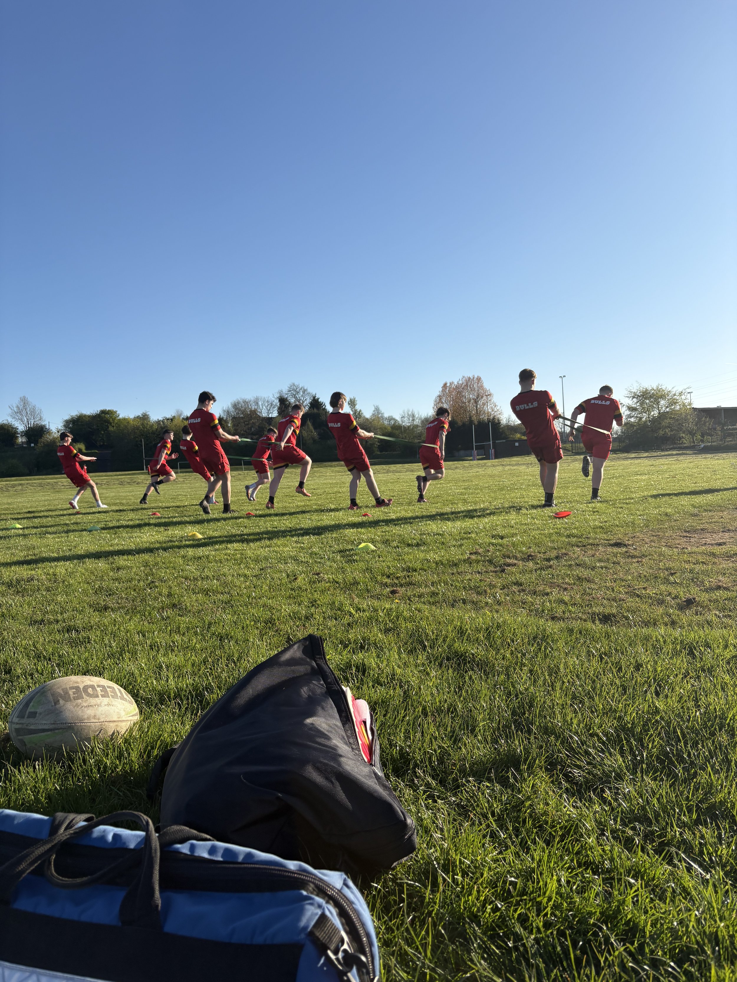 A group of young athletes in red sports uniforms running in a line on a grassy sports field during a sunny day, with trees in the background.
