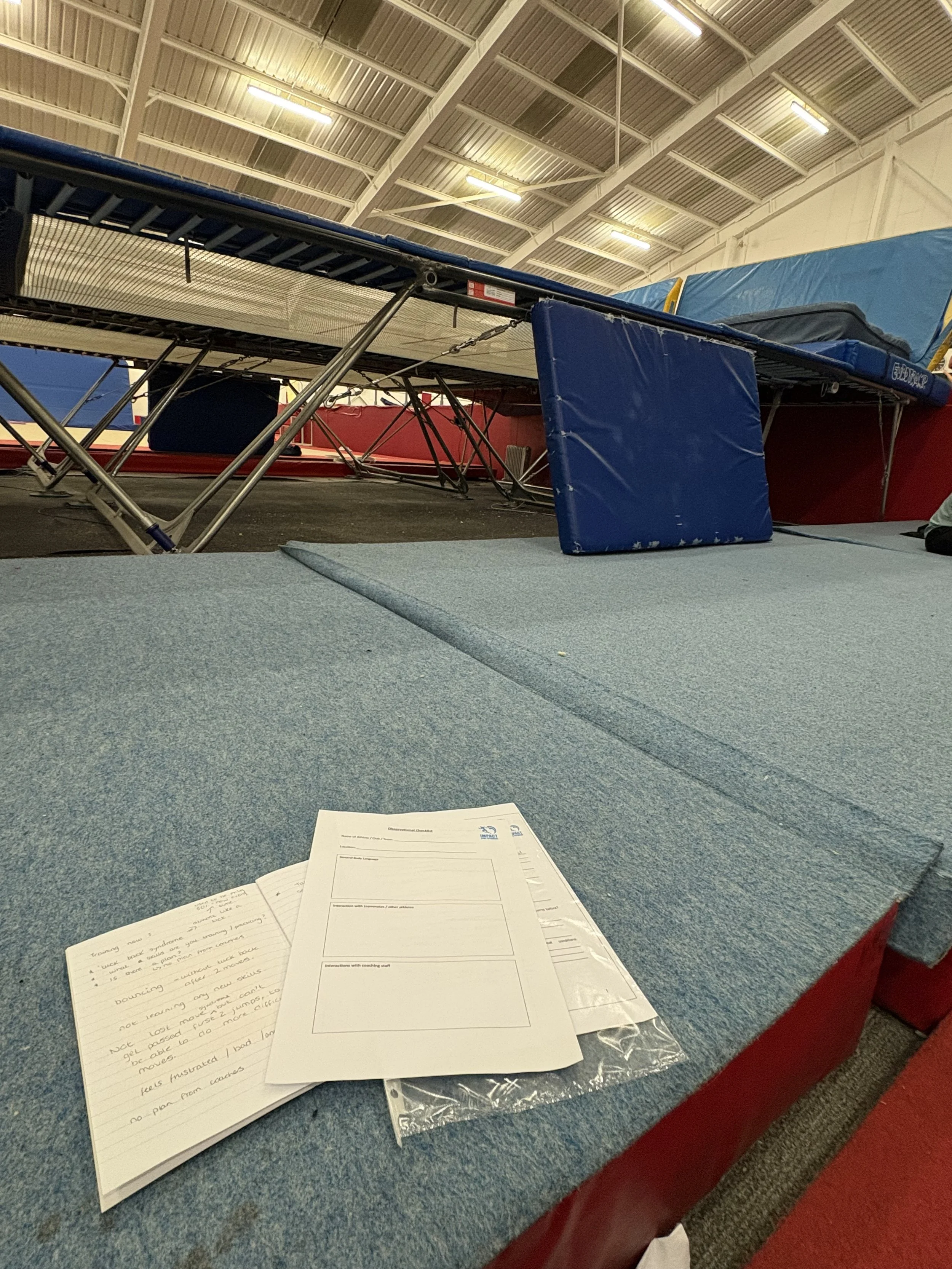 A table with handwritten notes, worksheets, and plastic sleeves on a blue tablecloth in a gymnasium or sports hall with a high ceiling and bright lighting.