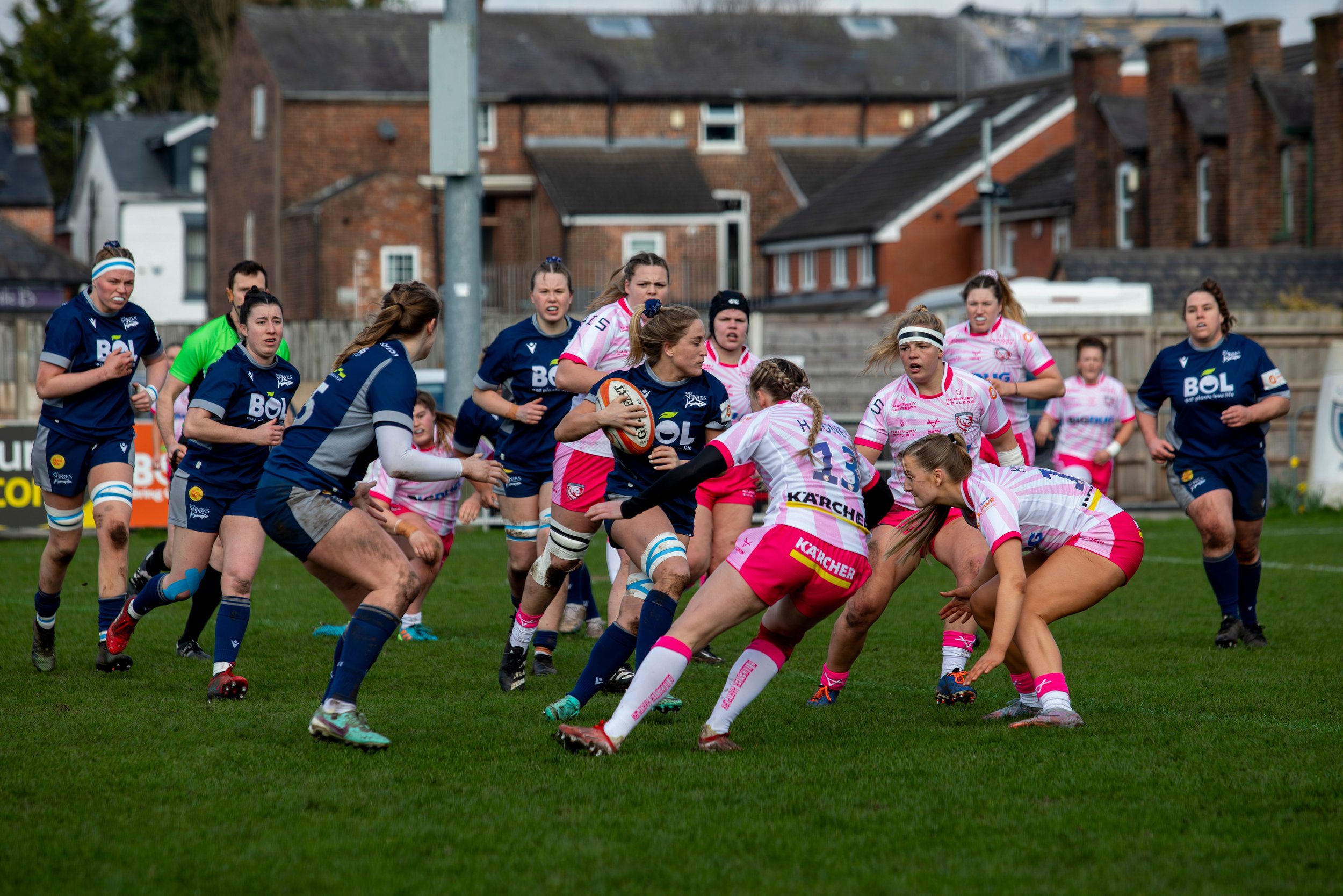 Women playing rugby on a grassy field during daytime, some wearing pink and white uniforms, others in navy blue, with houses in the background.