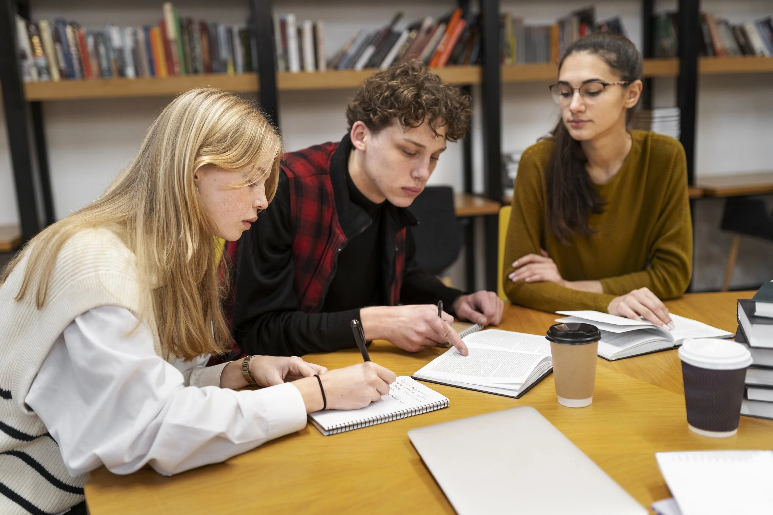 Three young adults sitting at a table studying together with books, notebooks, and coffee cups.