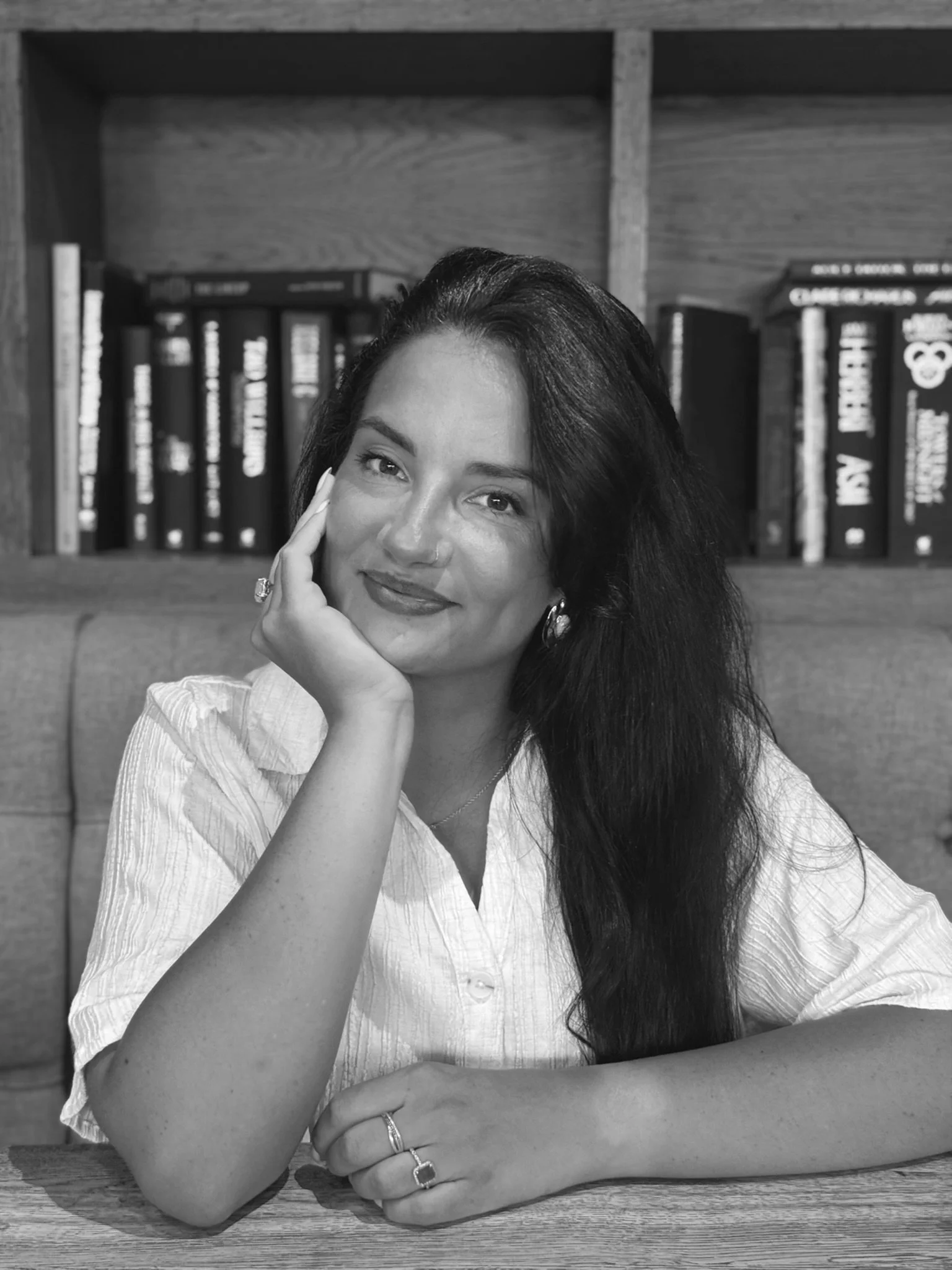 Black and white photo of a woman with long dark hair, resting her head on her hand and smiling, seated at a wooden table in front of a bookshelf filled with books.