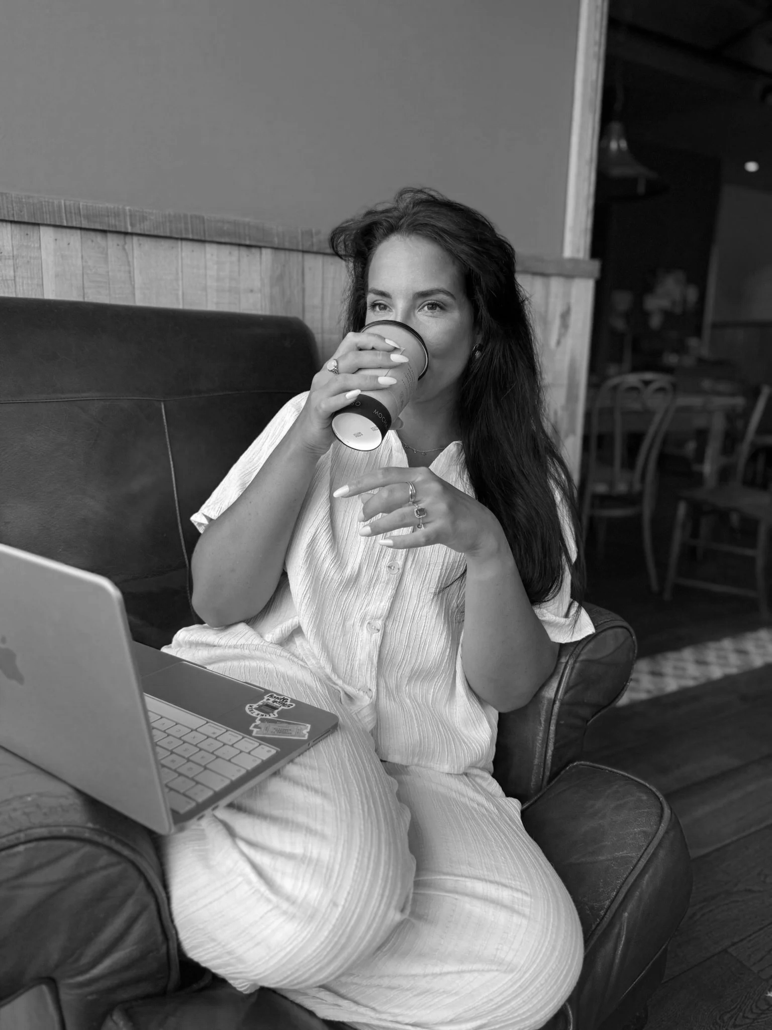 A woman with long dark hair sitting in a cafe or restaurant, drinking from a paper cup, with a laptop and phone in front of her, smiling and looking off-camera.