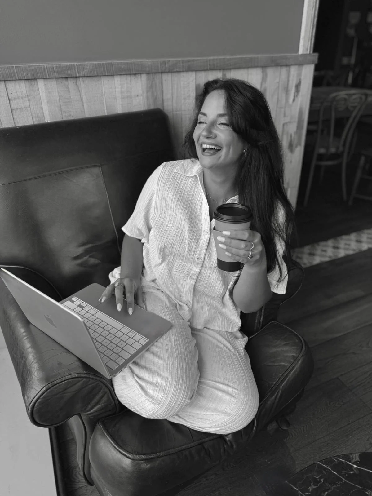 A woman with long dark hair, smiling and laughing, sitting on a leather couch in a coffee shop, holding a disposable coffee cup in one hand and using a laptop on her lap with the other hand.
