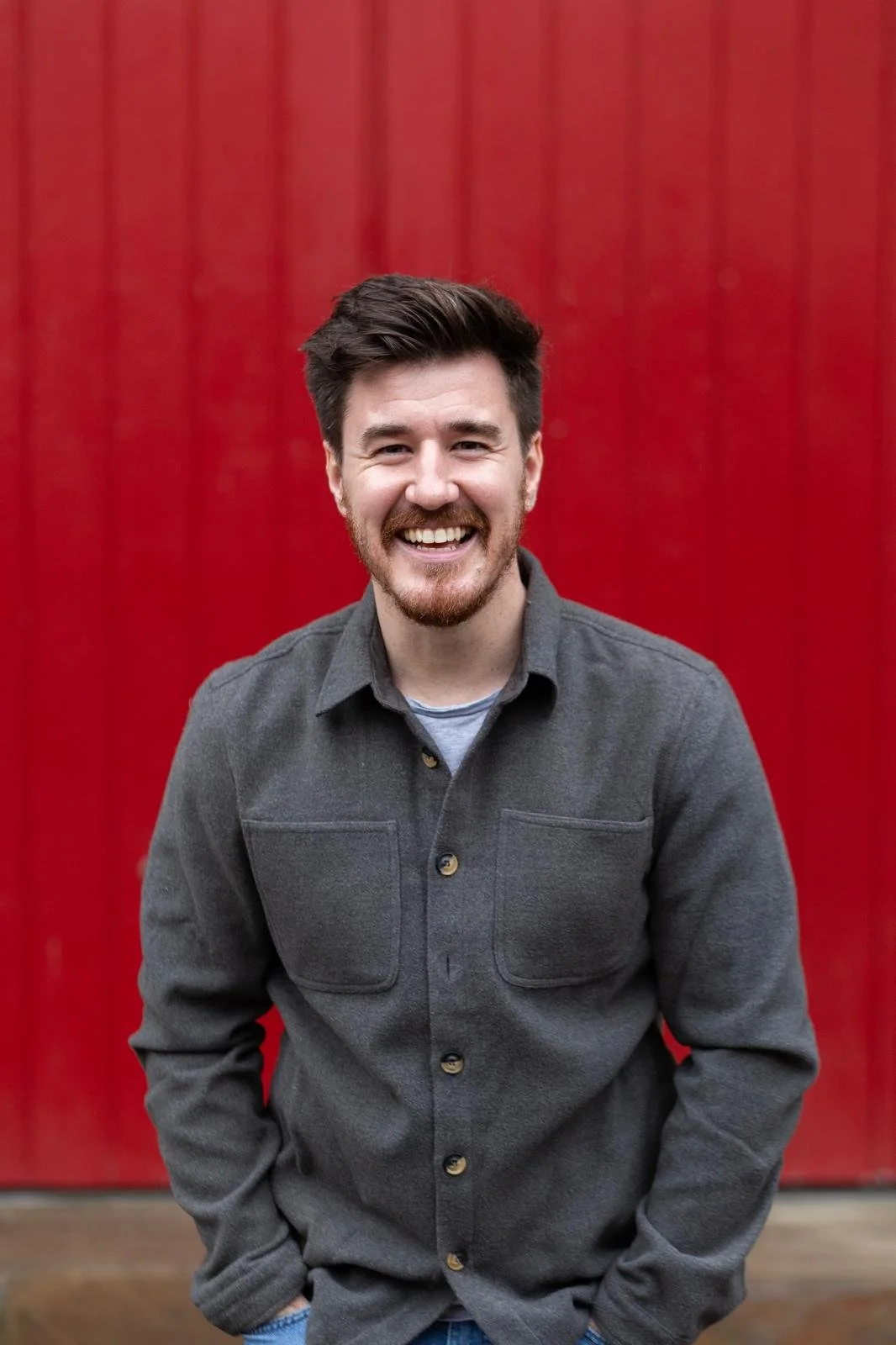 A smiling young man with dark hair and a beard, wearing a dark gray button-up shirt, standing in front of a red wooden wall.