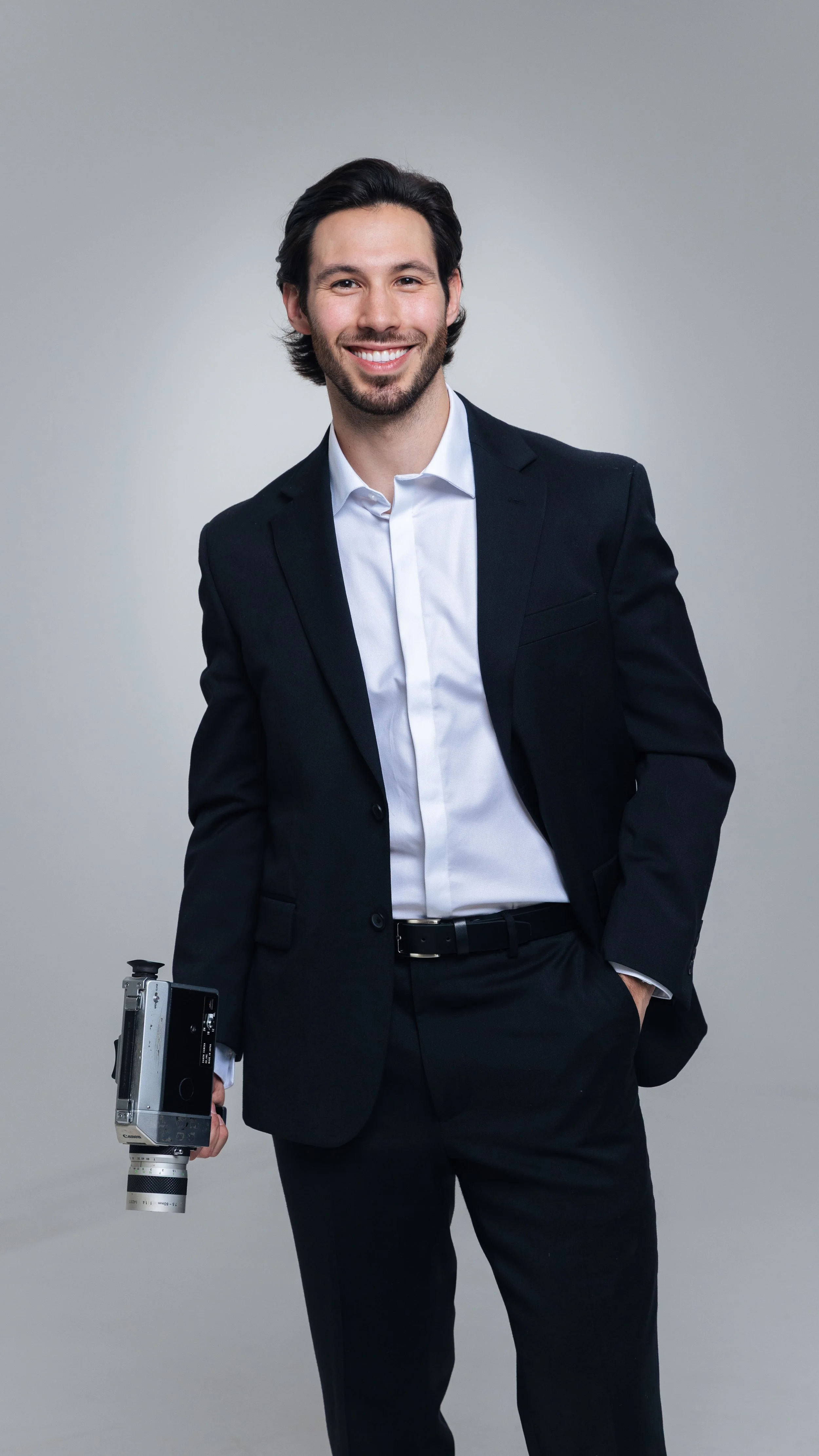 A man in a black suit holding a vintage camera, smiling, standing against a plain gray background.