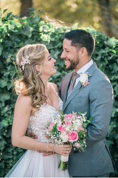 A newlywed couple in wedding attire standing outdoors with greenery in the background, looking at each other, holding a bouquet of pink and white flowers.