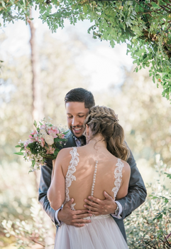 A groom and bride share a hug outdoors, with the groom holding a bouquet of pink and white flowers, celebrating their wedding under green foliage.