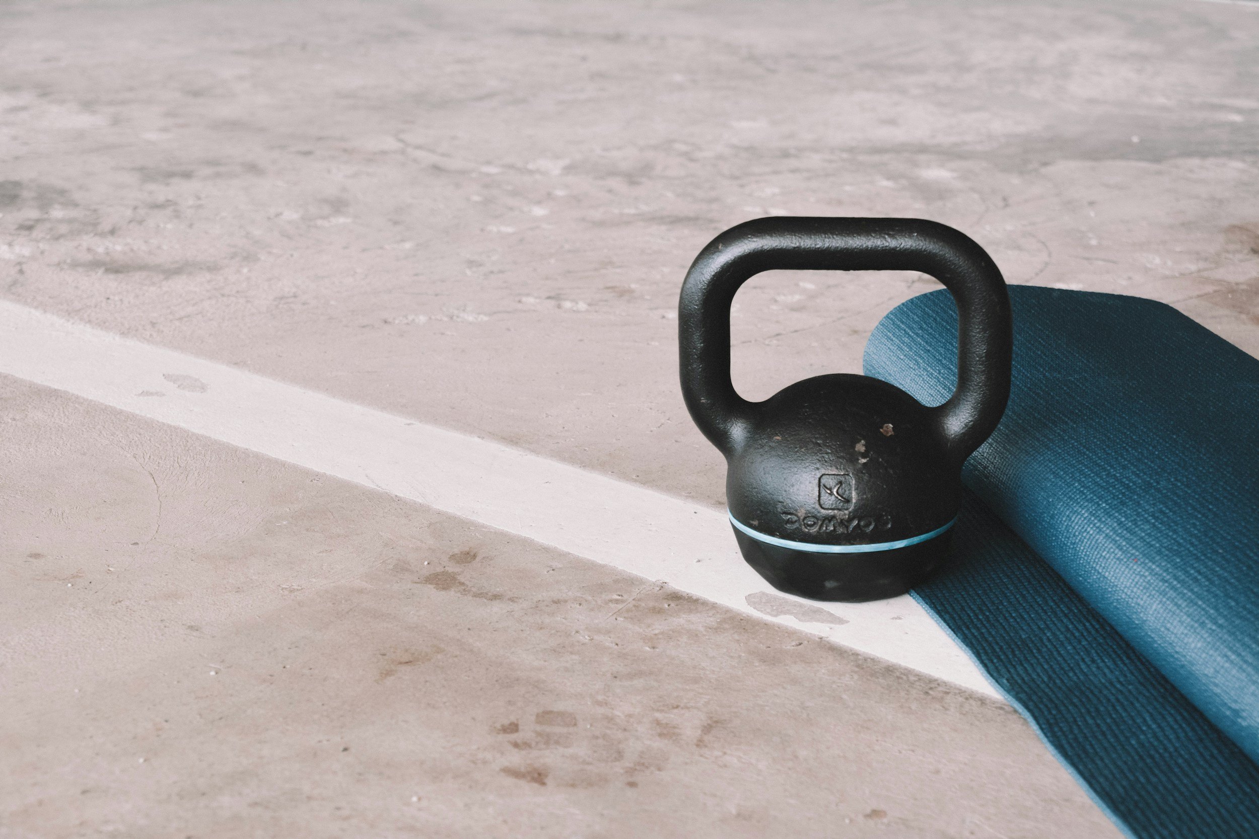 A black kettlebell resting on a gym mat next to a rolled-up blue exercise mat on a concrete floor.