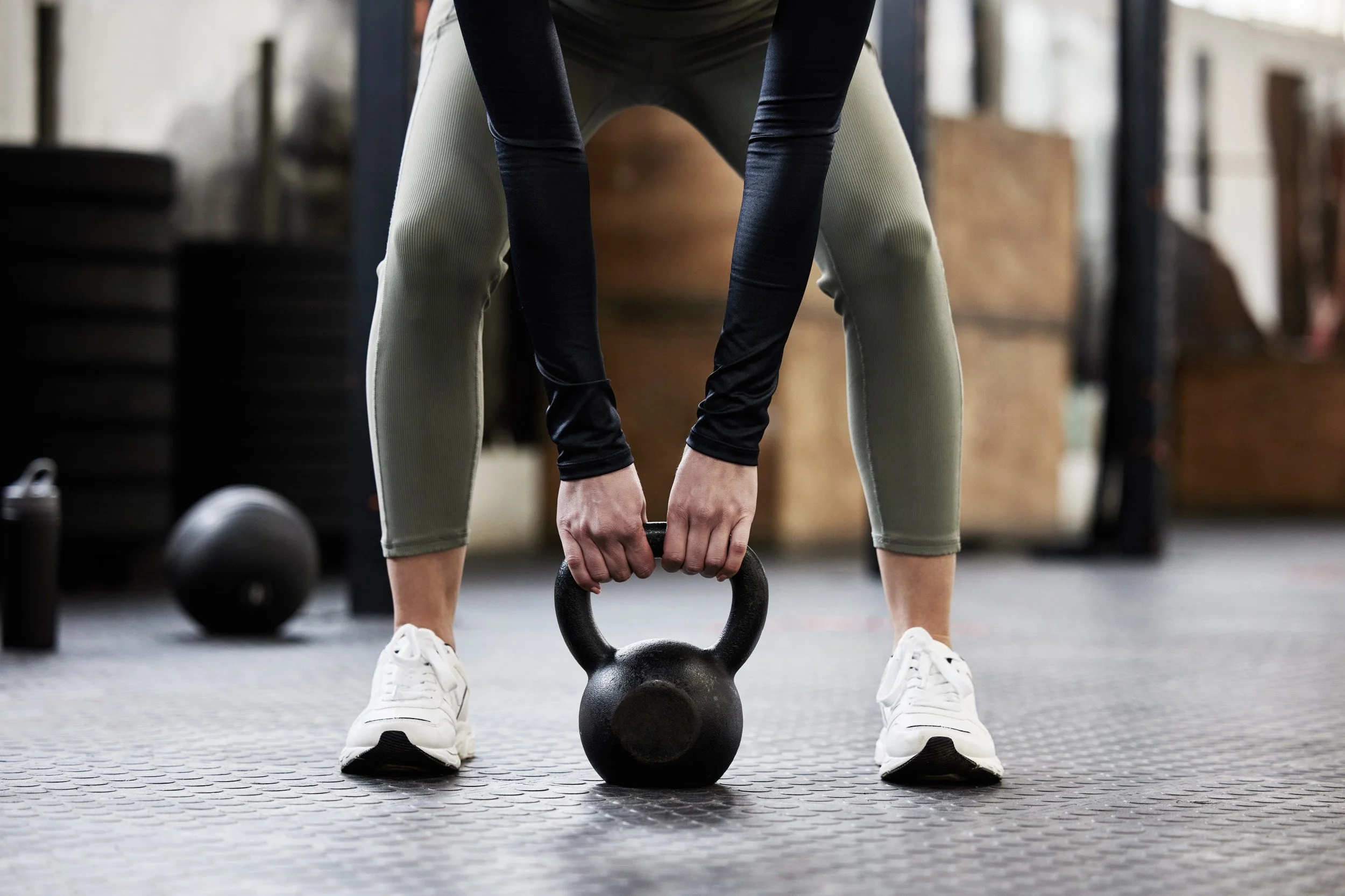 Person wearing gray leggings and white sneakers, holding a kettlebell in a gym.