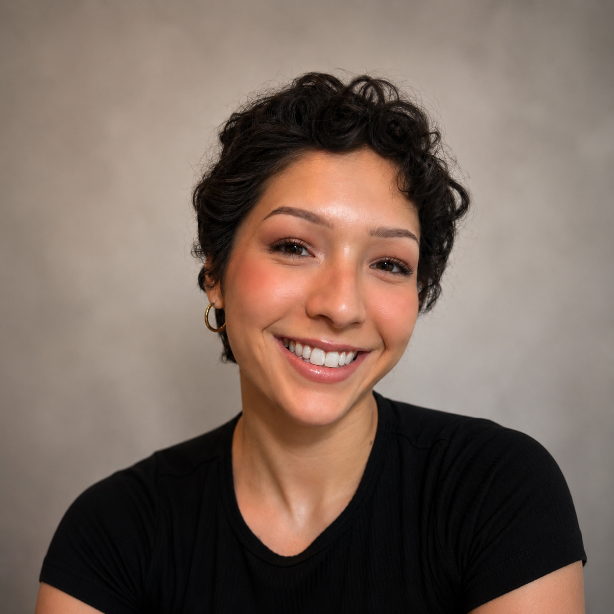 Close-up of a smiling woman with short wavy dark hair, wearing a black shirt and gold hoop earrings, against a plain neutral background.