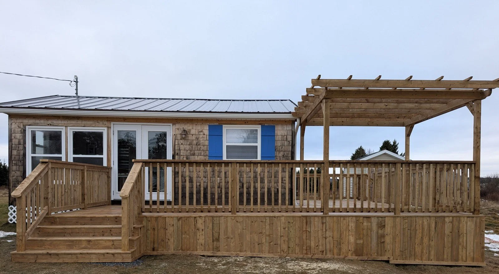 Custom built, pressure treated, wooden wrap around deck with wood railings, pergola and stairs at each end, built for a cottage owner in Canoe Cove, PEI.