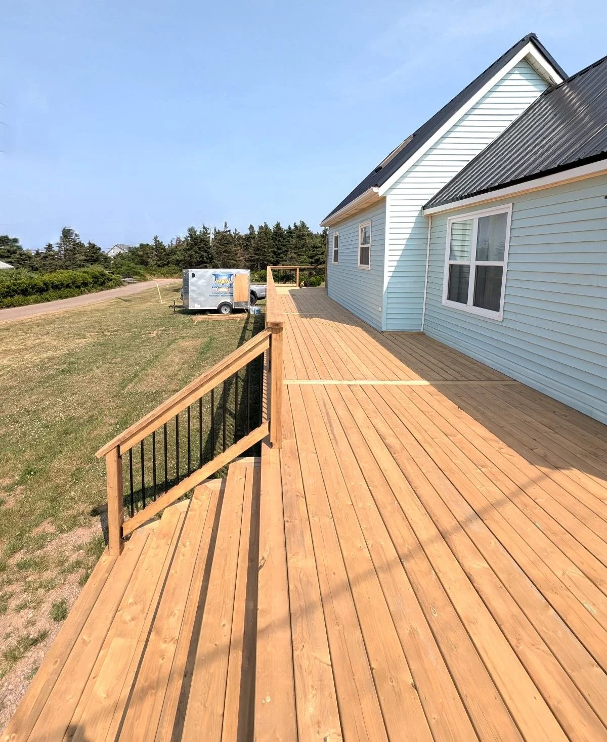 Custom built, pressure treated, wrap-around,  wooden deck with wood & black aluminum railings and 12-ft wide stairs, built for a home owner in Wild Harbour, PEI