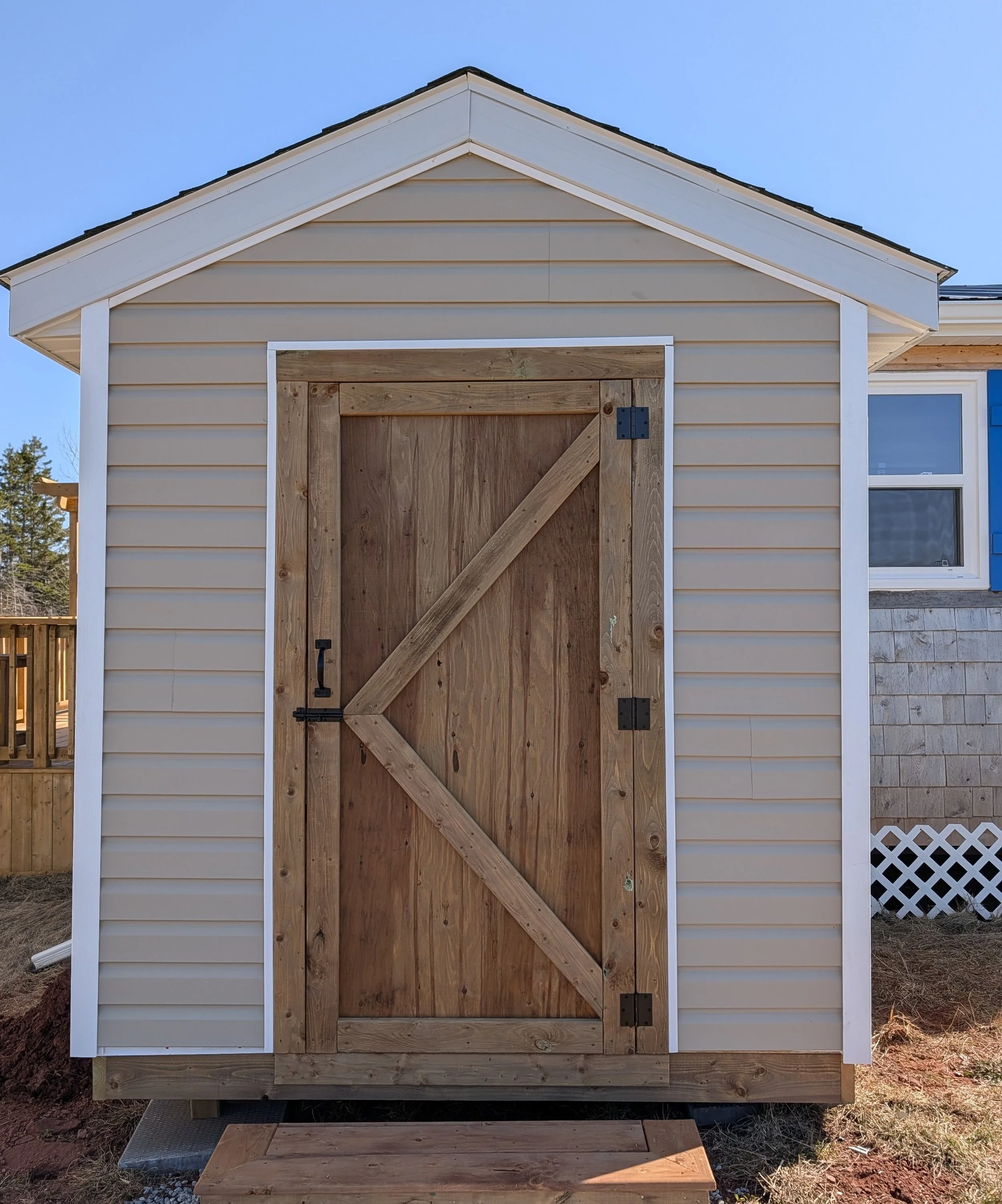 Custom designed and built detached storage shed with vinyl siding and wooden door built for cottage owner in Canoe Cove, PEI.