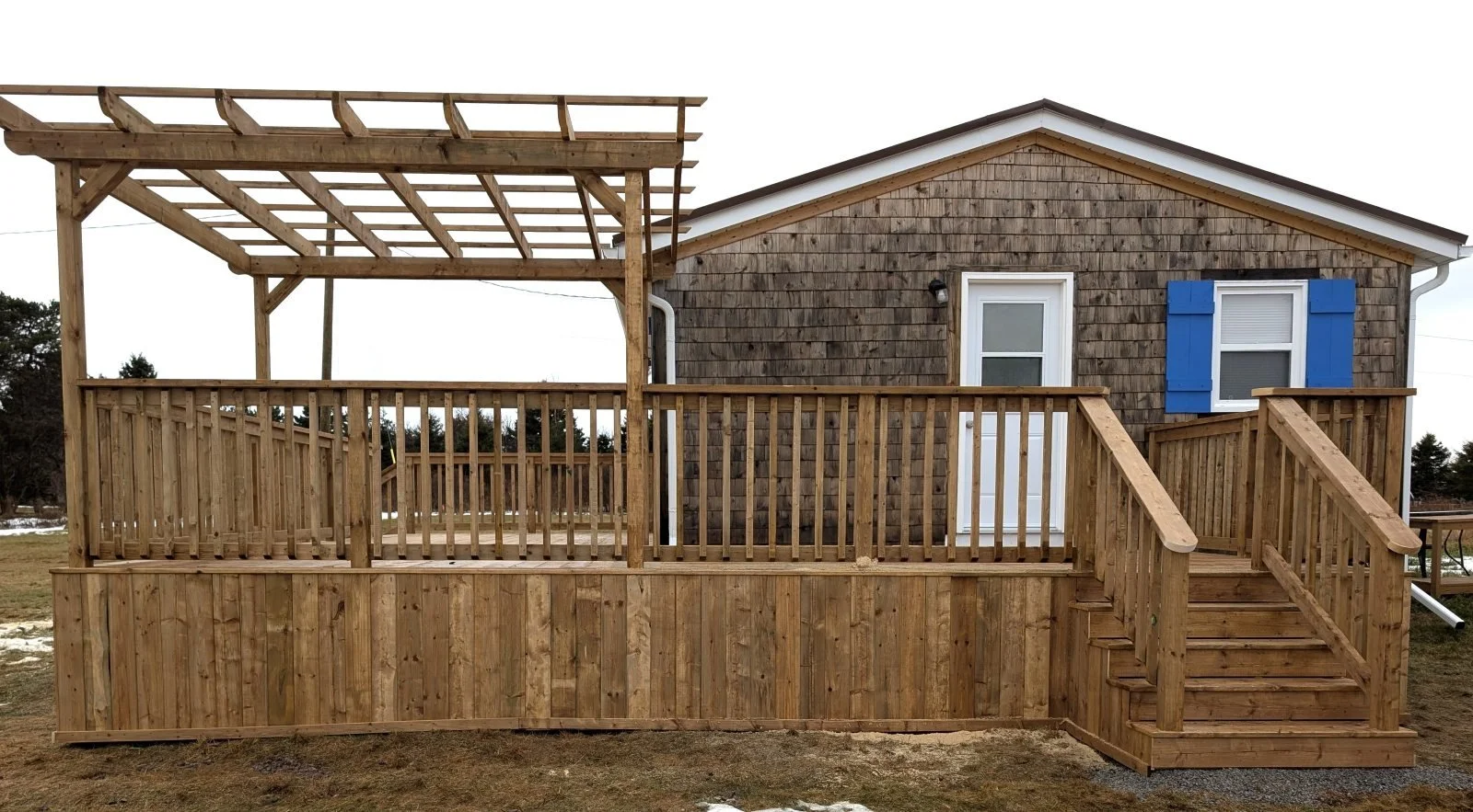 Custom built, pressure treated, wooden wrap around deck with wood railings, pergola and stairs at each end, built for a cottage owner in Canoe Cove, PEI.