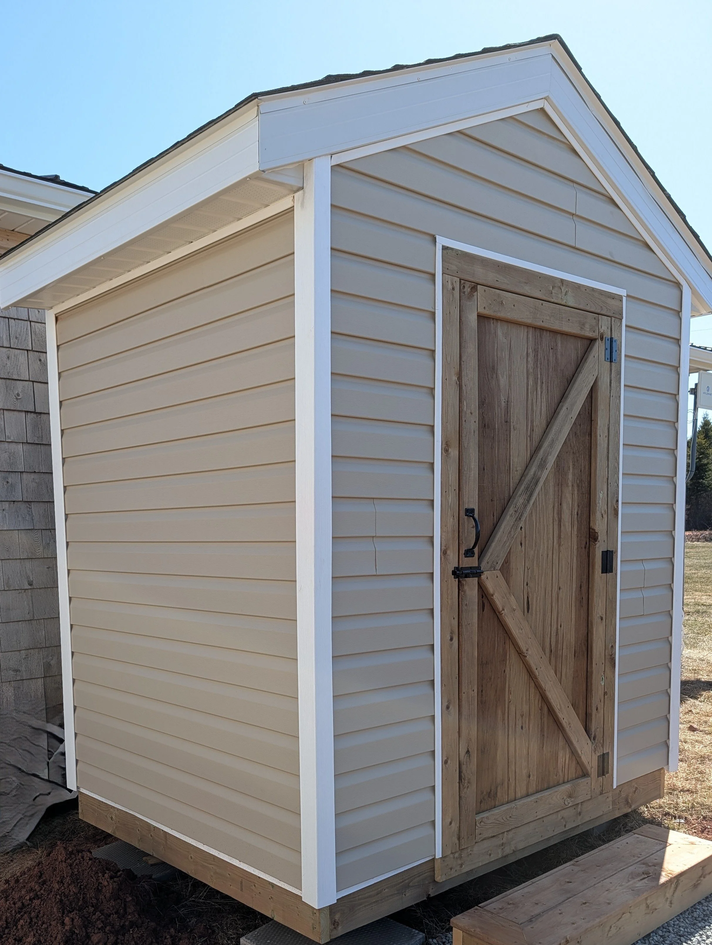 Custom designed and built detached storage shed with vinyl siding and wooden door built for cottage owner in Canoe Cove, PEI.