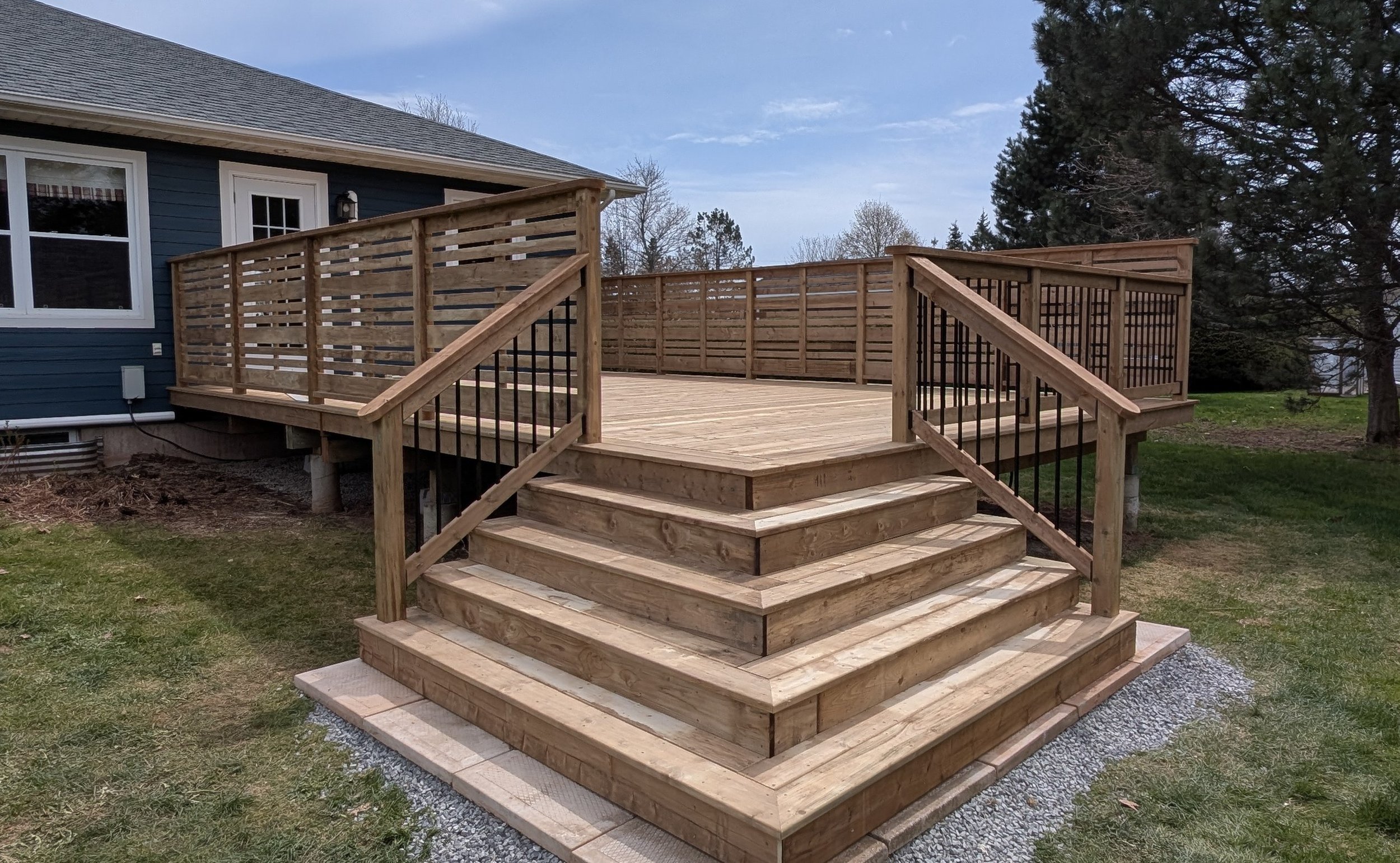 Completed pressure-treated wooden deck with privacy walls, black aluminum railings and cascading stairs built in Winsloe, Prince Edward Island