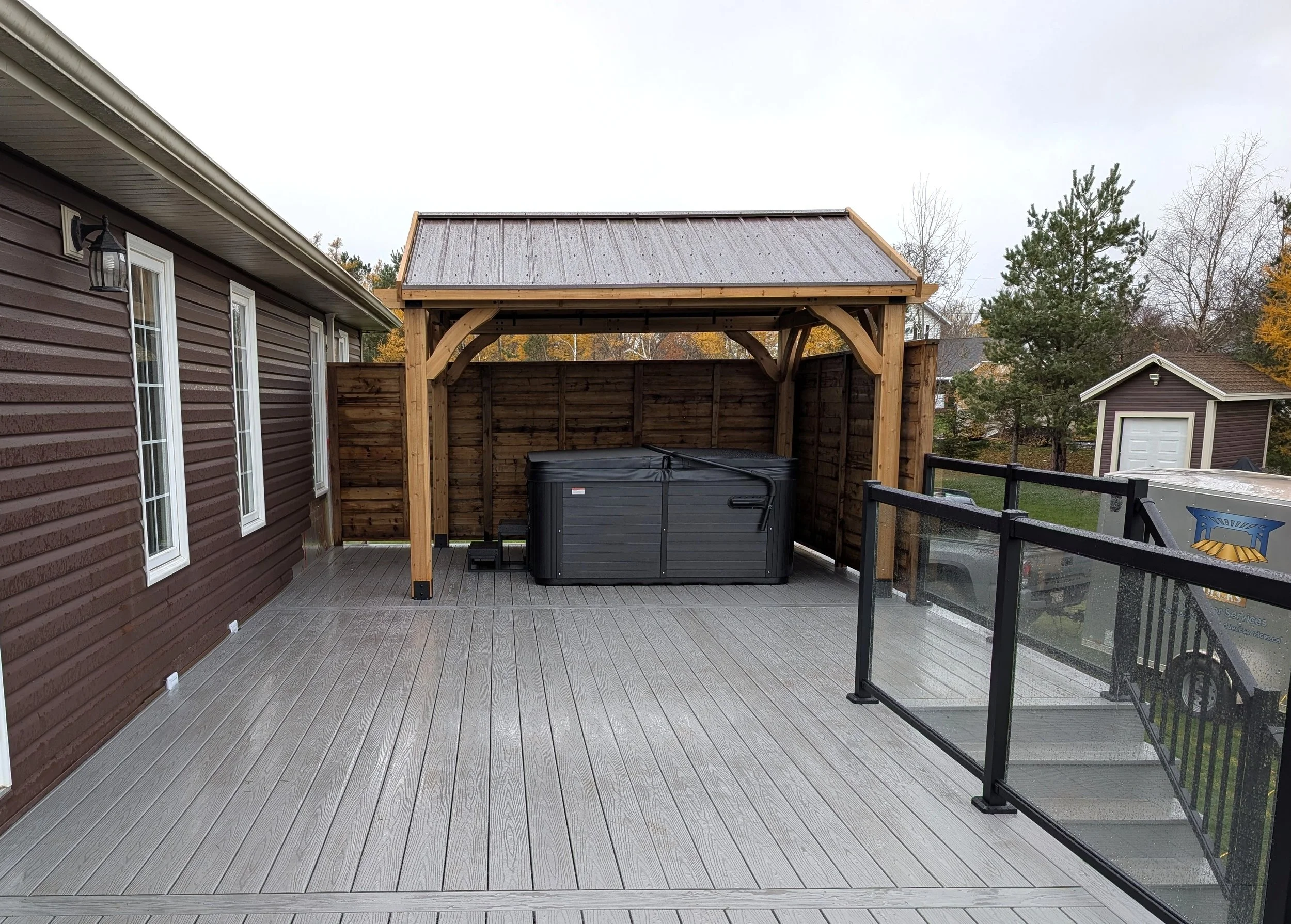 Wood pergola hot tub enclosure with metal roof on composite deck with glass & black aluminum railings built for a homeowner in Cornwall, PEI 