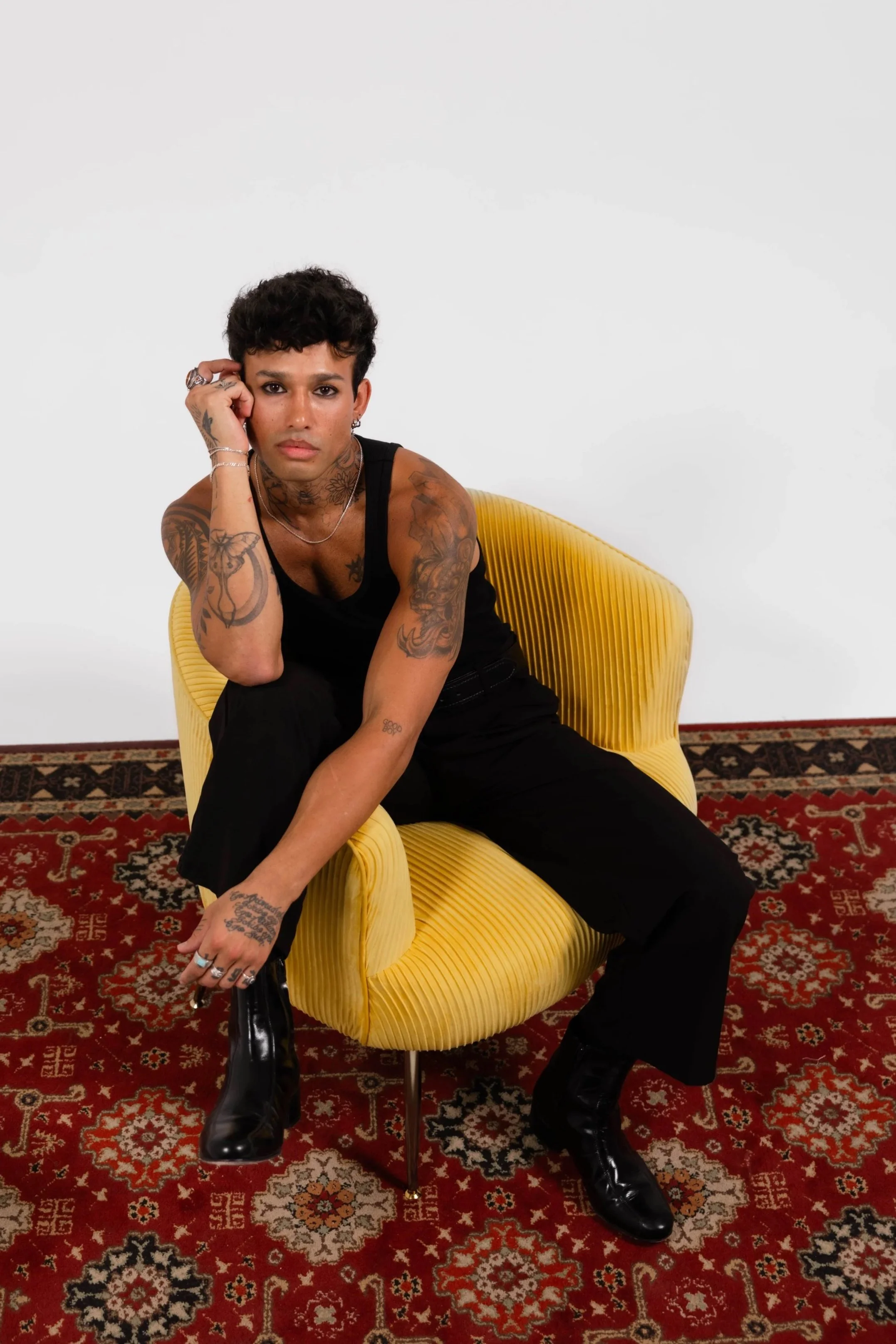 A young person with short dark hair, tattoos on arms and neck, wearing a black sleeveless top and black boots, sitting on a yellow modern chair with a curved design, against a plain white wall, on a patterned red carpet.