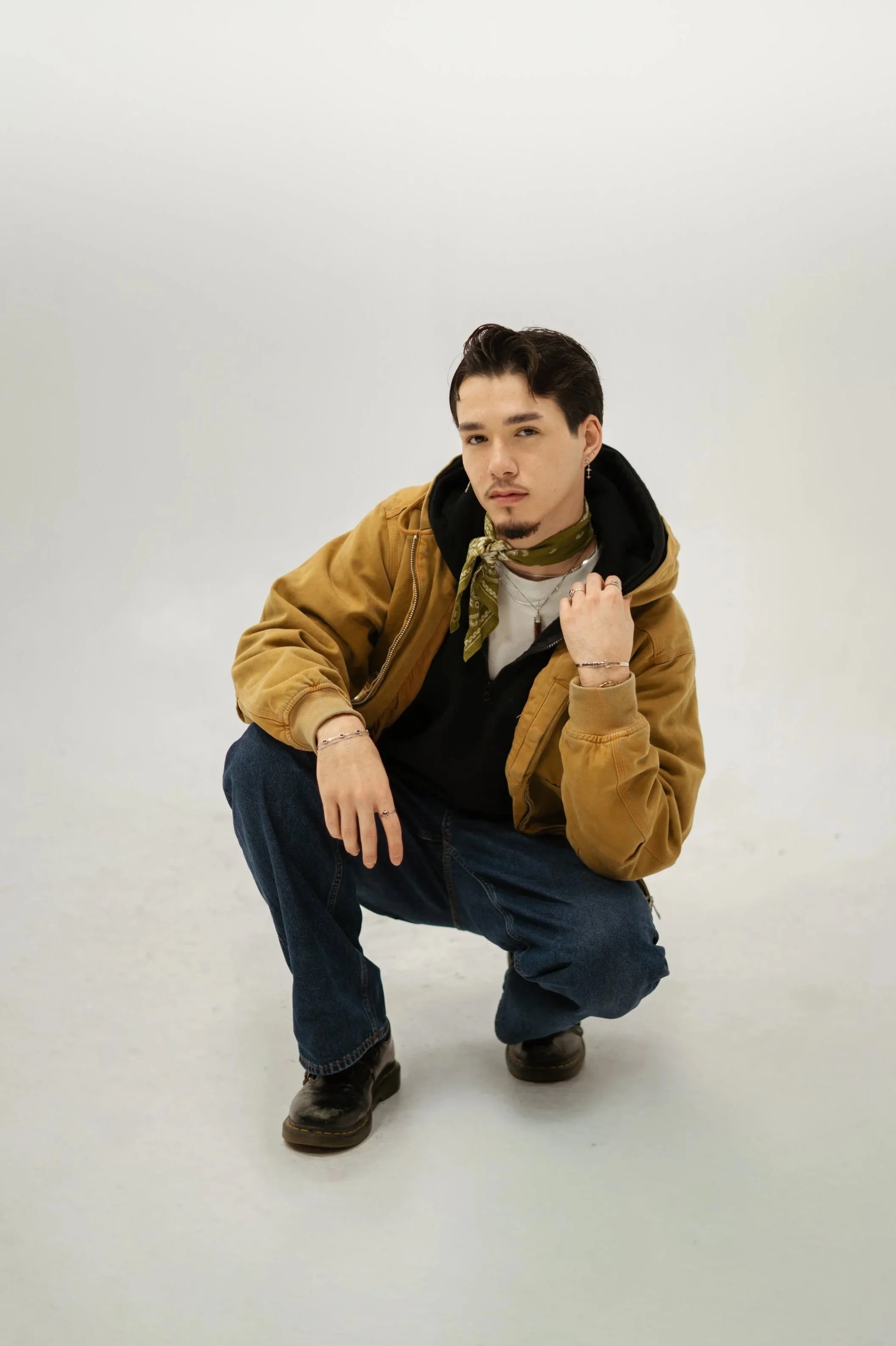 A young man crouching in front of a plain light-colored background, wearing a tan jacket, black hoodie, jeans, and boots, with a green bandana around his neck, holding his collar with one hand and resting his other hand on his knee.