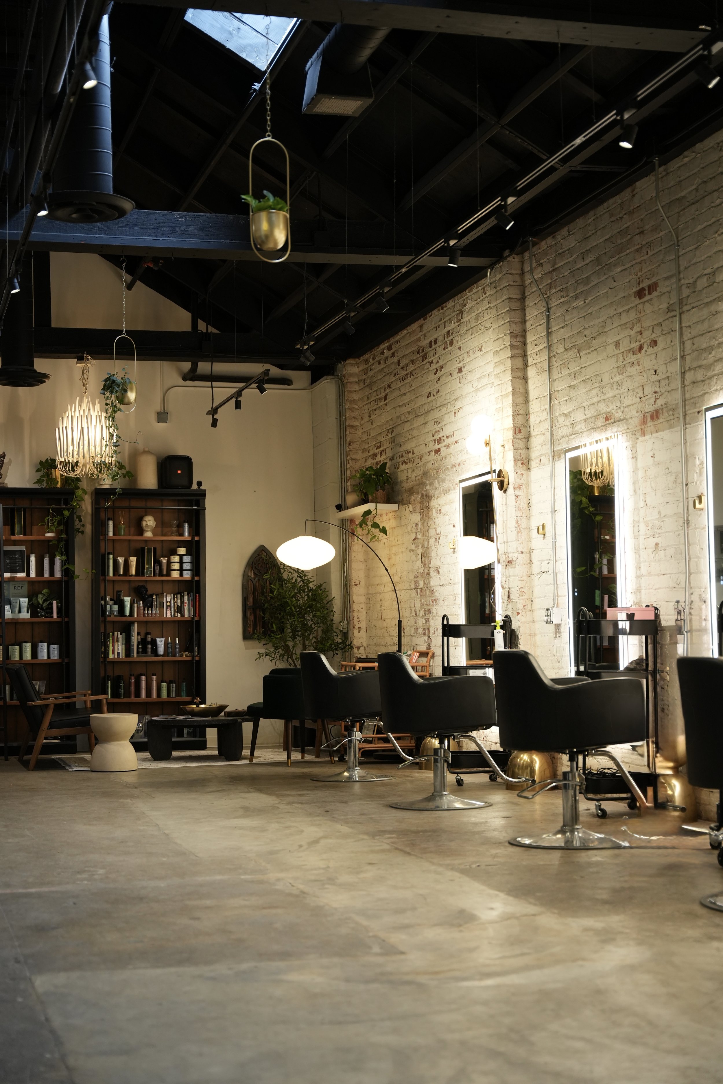 Interior of a modern salon with multiple black barber chairs, a brick wall with mirrors, and decorative plants and lighting fixtures.