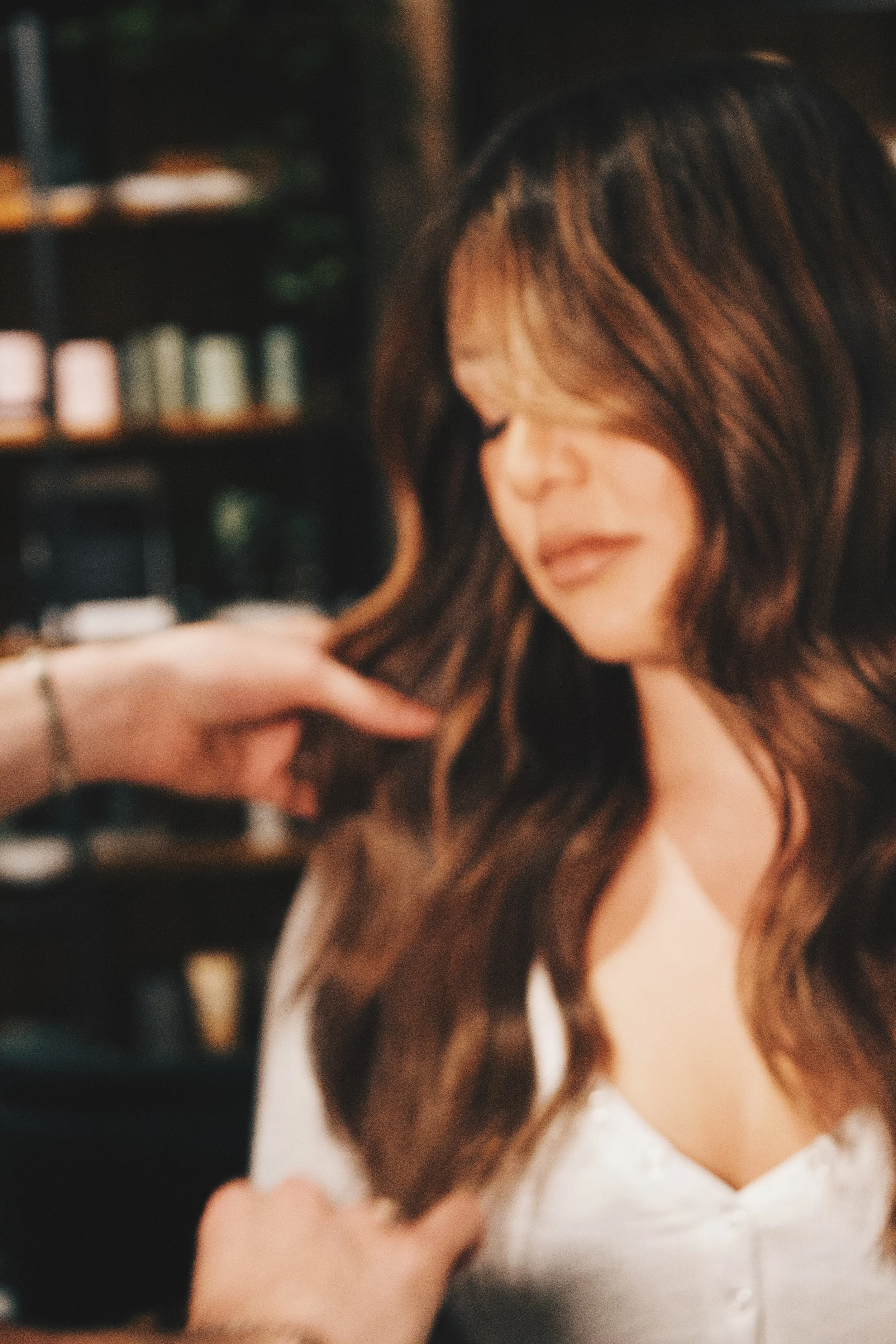A woman with long, wavy brown hair is getting a hair styling or hair coloring treatment in a salon, with shelves and hair products in the background.
