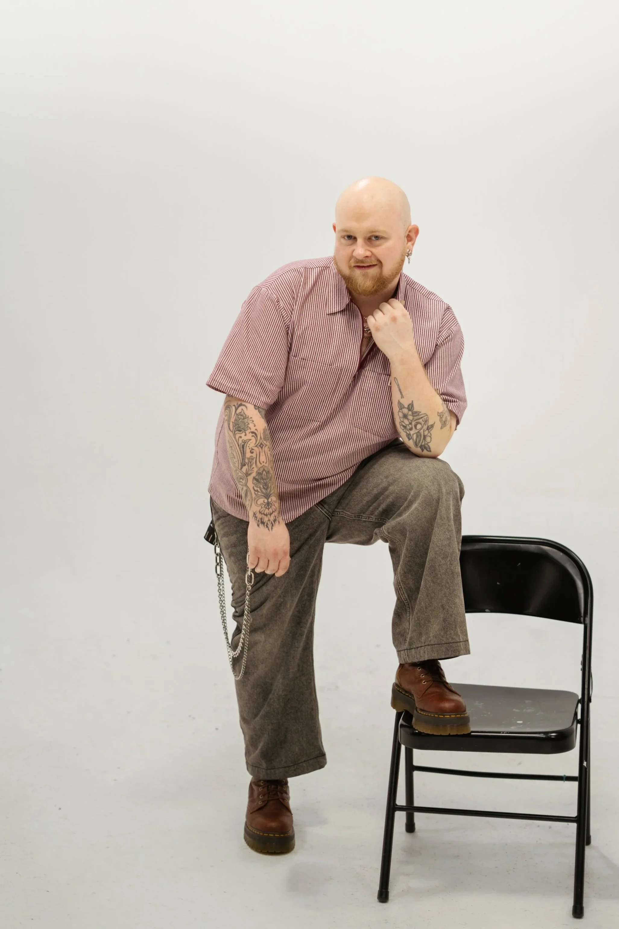 A man with tattoos, wearing a striped shirt, brown boots, and gray pants, posing with one foot on a black chair in a white studio environment.